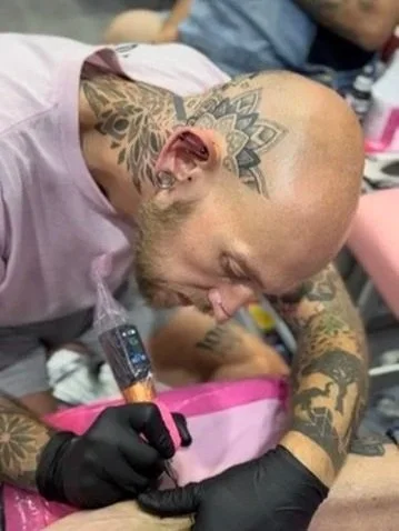A tattooed man with a shaved head and earrings getting a tattoo on his arm, wearing black gloves and a pink shirt.