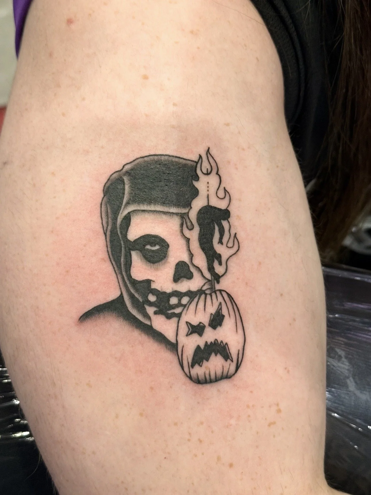 Tattoo of a skull with dark shading, a third eye on the forehead, and a pumpkin with a carved face hanging below it, with flames coming out of the skull's head.