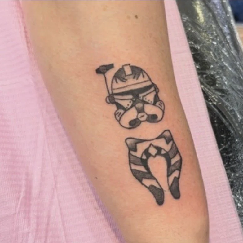 Tattoo of a cartoon-style stormtrooper from Star Wars with a small cat, both upside down on a person's forearm.