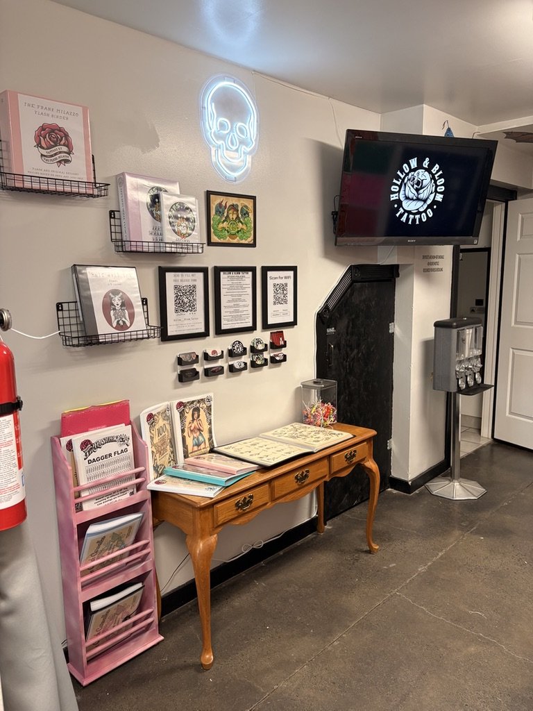 Interior of a tattoo studio with a neon skull sign on the wall, framed artwork, QR codes, tattoo aftercare products, a small wooden table displaying tattoo design books, and a large monitor screen showing the studio's logo and name 'Hollow & Blood Ta