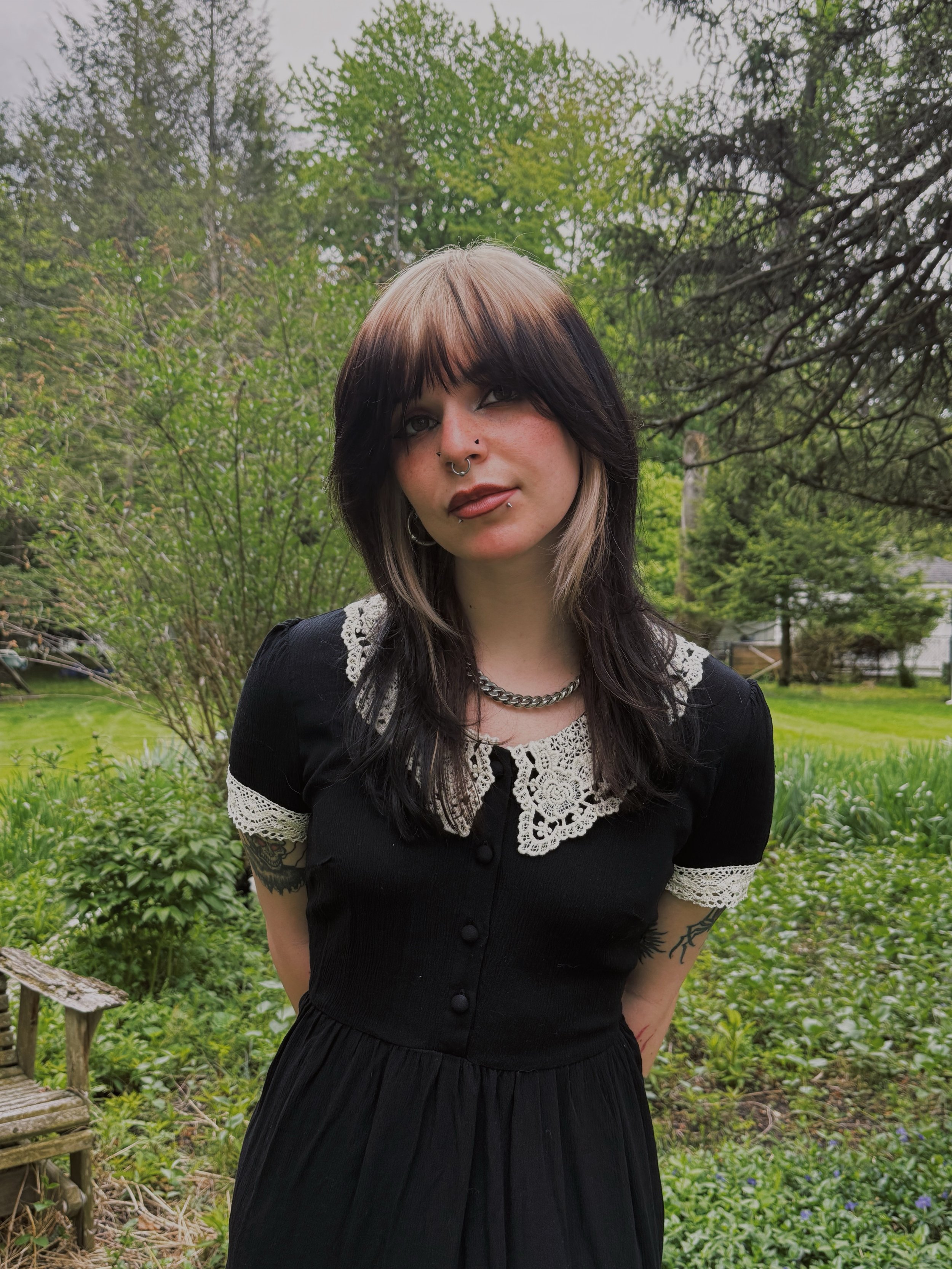 A woman with dark and blond hair wearing a black dress with lace details standing outdoors in a garden.