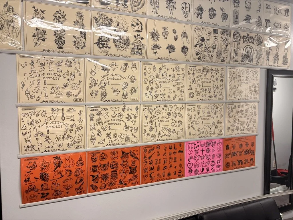 Display of tattoo flash sheets with black ink tattoo designs organized by theme and color, including traditional, Halloween, and pink-themed sheets.