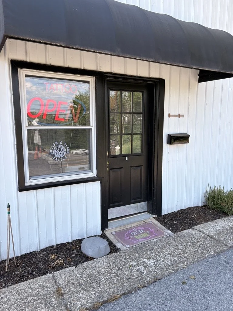 Outside entrance of a building with a black door and white siding, A red neon "Open" sign and a pink "Tattoo" sign in the window, a black mailbox, a welcome mat, and a small bush.