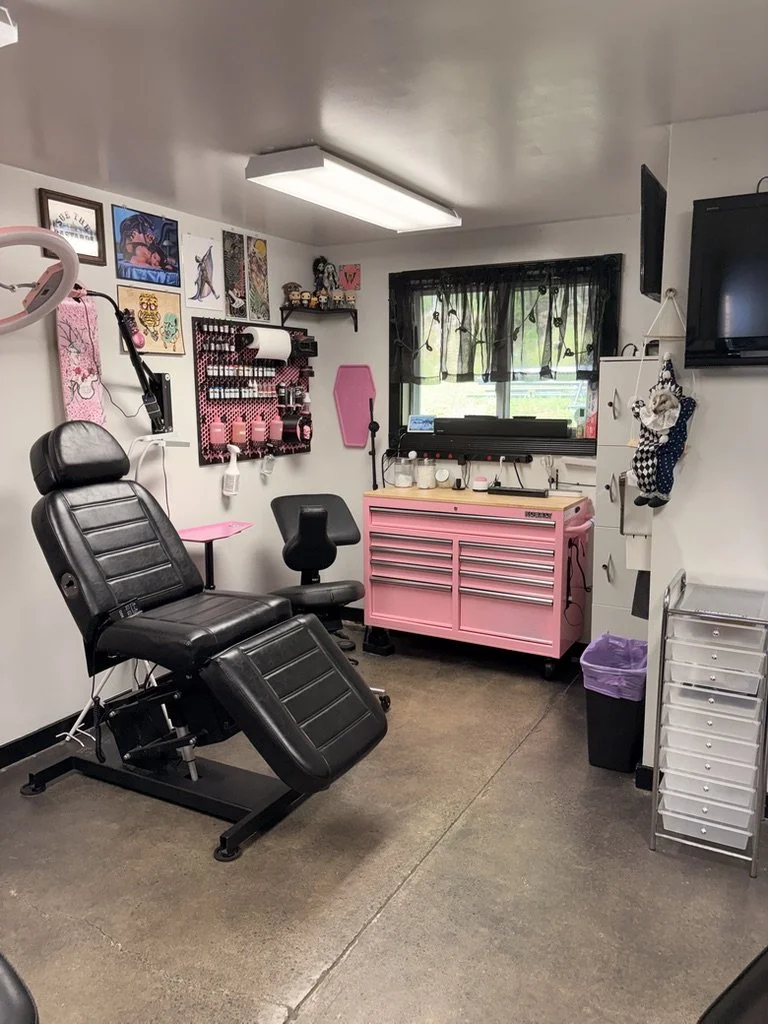Tattoo studio with tattoo chair, wall art, pegboard with supplies, pink work table, and equipment for tattooing.