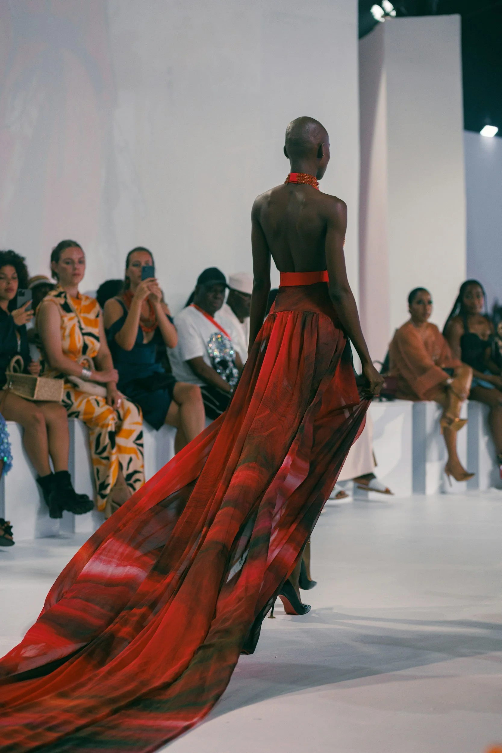 A fashion model walks on a runway wearing a flowing red and green patterned dress, with an audience seated on either side watching and taking photos.