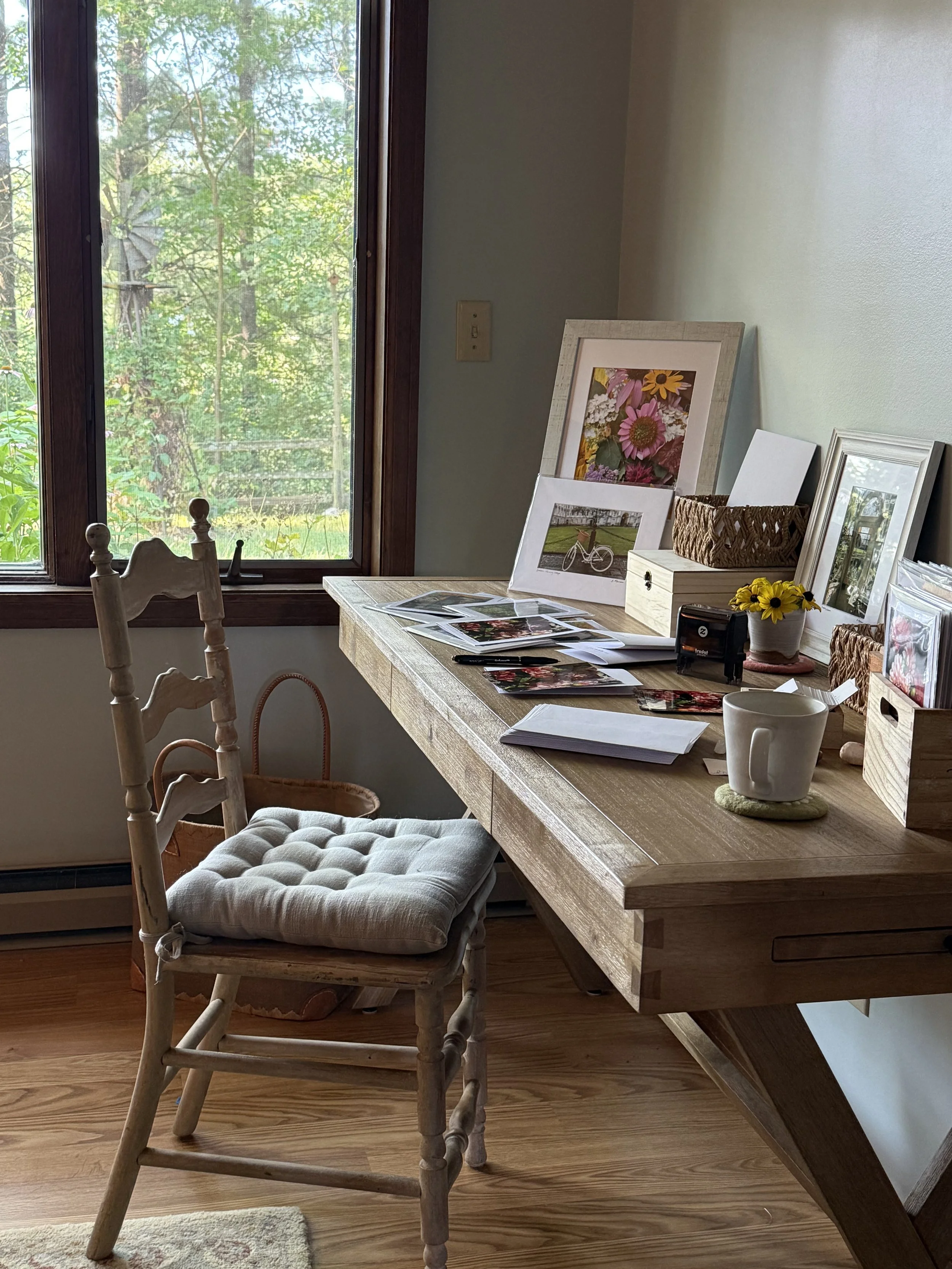 Indoor scene showing a wooden dining table with a cushioned armchair, a window overlooking trees, framed pictures, photos, a small potted flower, and some documents.