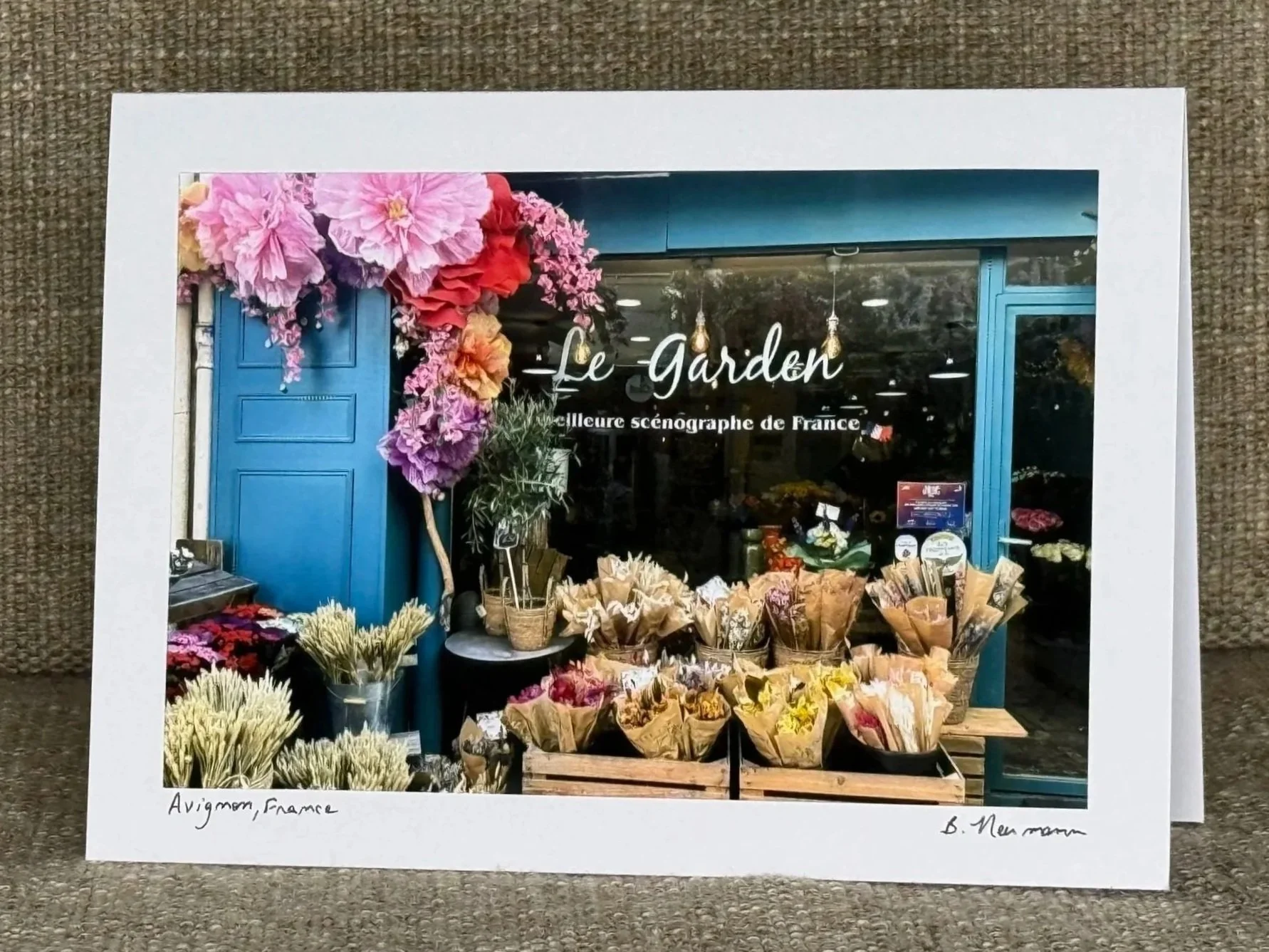 Photo of a storefront with the sign 'Le Garden' and flowers outside, including pink, purple, yellow, and orange blossoms in buckets and paper wraps.