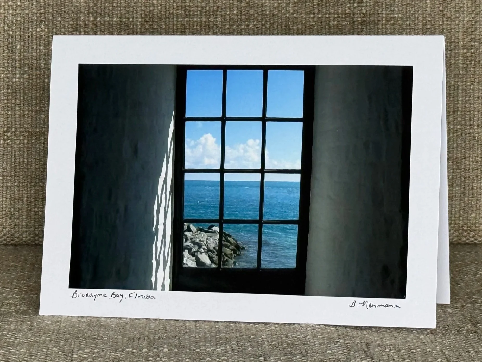 Photograph of a window overlooking the ocean with rocks below and a blue sky with clouds, taken in Biscayne Bay, Florida.
