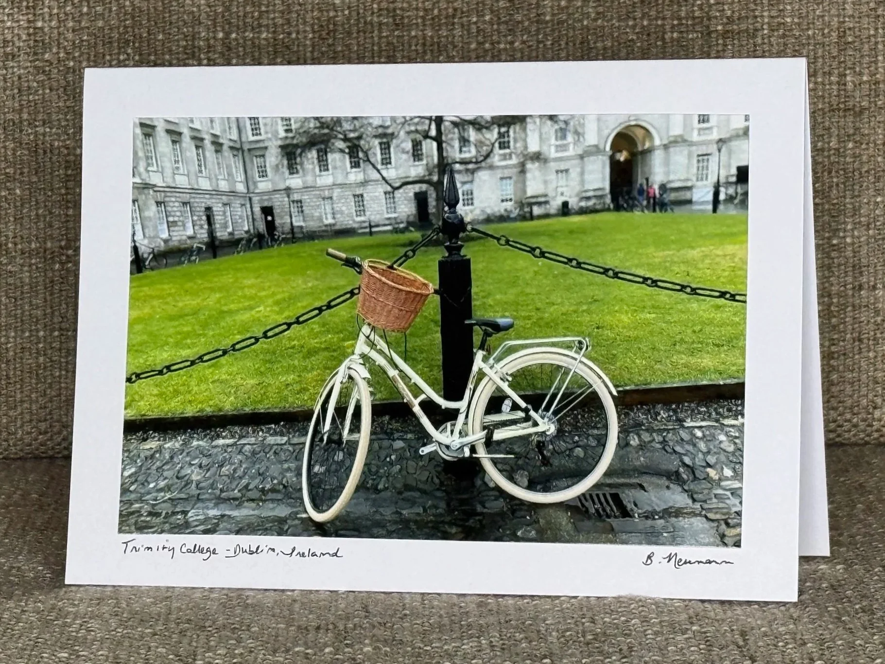 Photograph of a white bicycle with a wicker basket, parked and chained to a black post, in front of Trinity College Dublin, Ireland. The background shows a historic stone building with a large arched entrance and a grassy area with few people. The ph