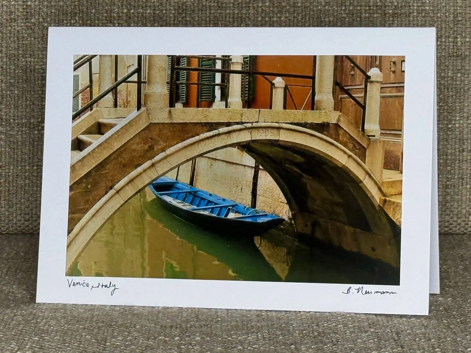 A printed photograph of a canal scene in Venice, Italy, featuring a small blue boat docked under a stone bridge with ornate railings and building facades in the background. The white border underneath the photo has handwritten text that says 'Venice 
