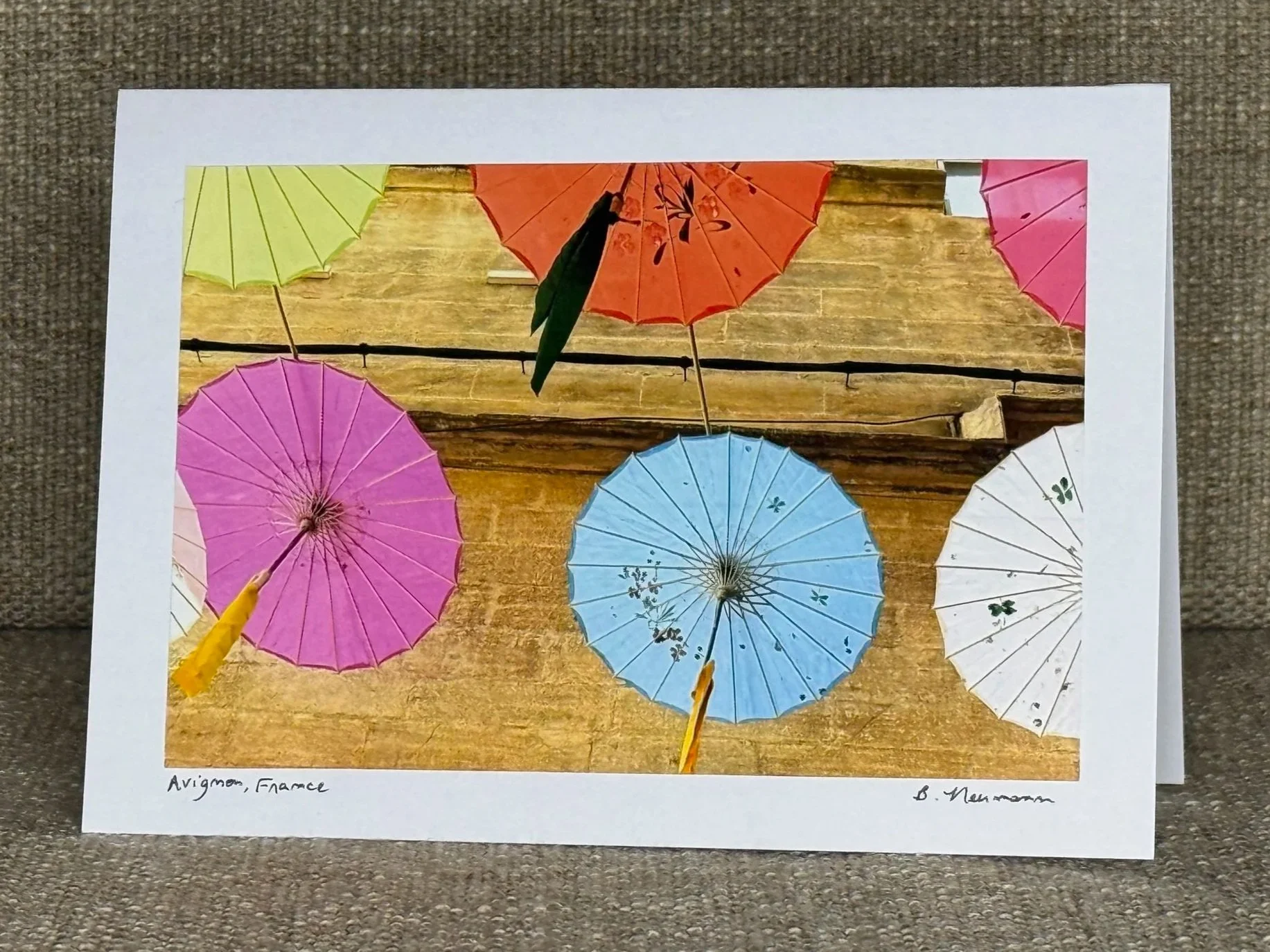 Photograph of colorful paper umbrellas with a wooden background, with a person in a yellow jacket holding a pink umbrella in the foreground. The photo is printed on a white-bordered sheet of paper placed on a textured surface. Handwritten text at the