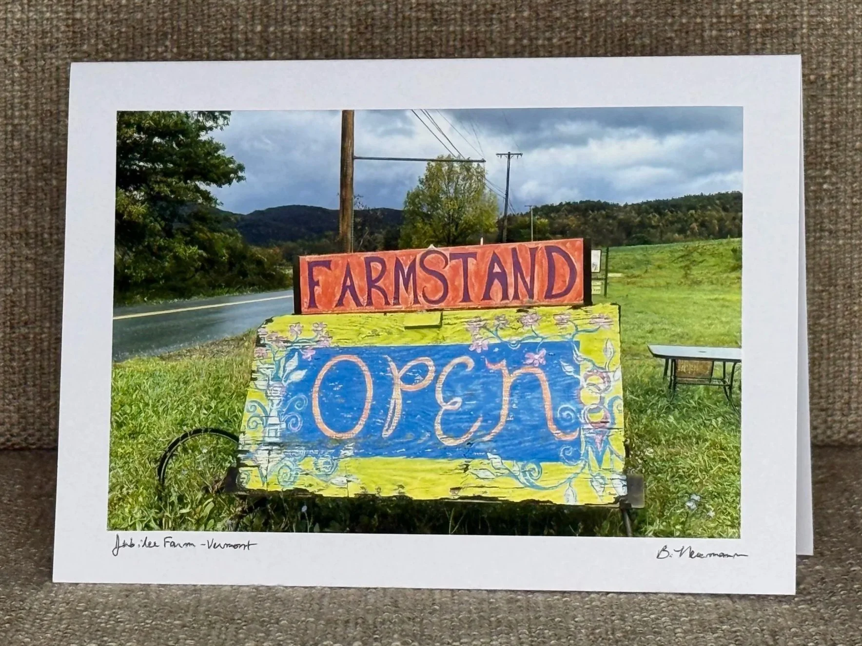 Colorful outdoor sign with the word 'OPEN' at the bottom and 'FARMSTAND' at the top, set against a rural landscape with hills, trees, power lines, and a cloudy sky.