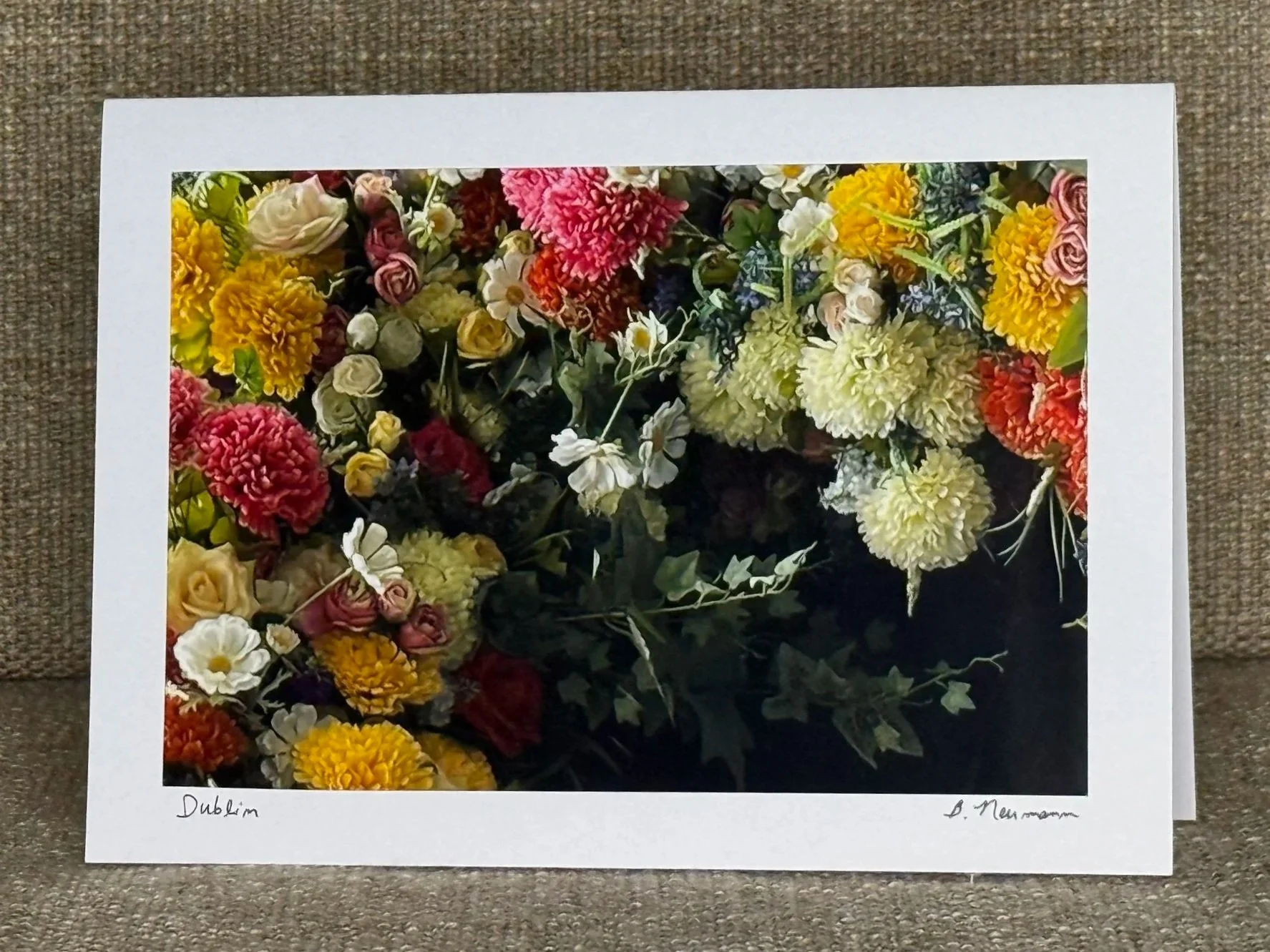 A framed photograph of a colorful flower arrangement with various flowers including roses, chrysanthemums, and daisies, placed on a brown textured surface.