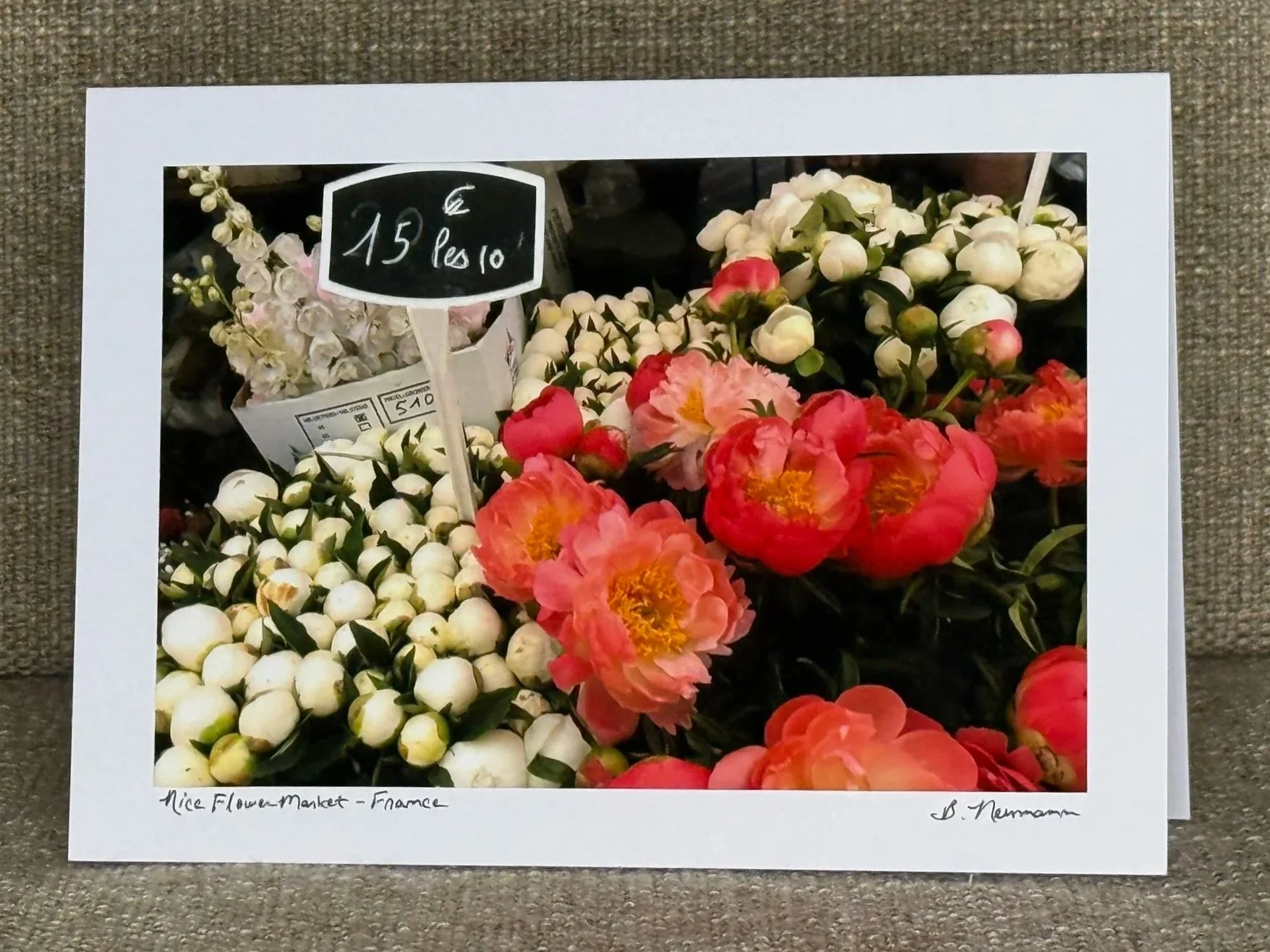 A framed photograph of flowers from Nice Flower Market in France. The photo shows white and pink flowers, with a small blackboard sign indicating a price of 15 euros for 10 flowers. Handwritten text at the bottom of the frame reads 'Nice Flower Marke