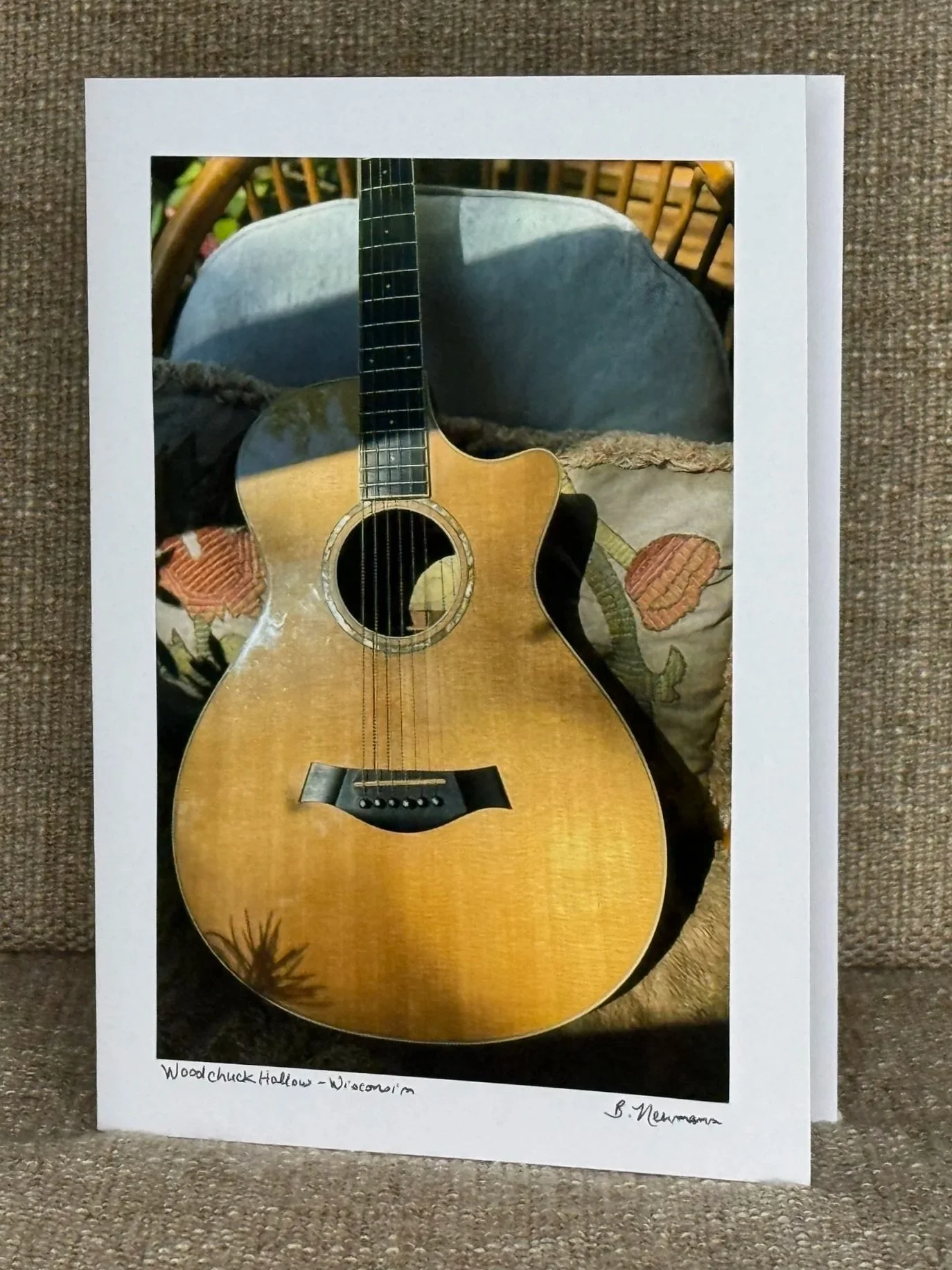 Photograph of a printed photograph of an acoustic guitar resting on a pillow with floral embroidery, placed on a cushioned surface. The printed photograph is surrounded by a white border with handwritten text at the bottom of the border.