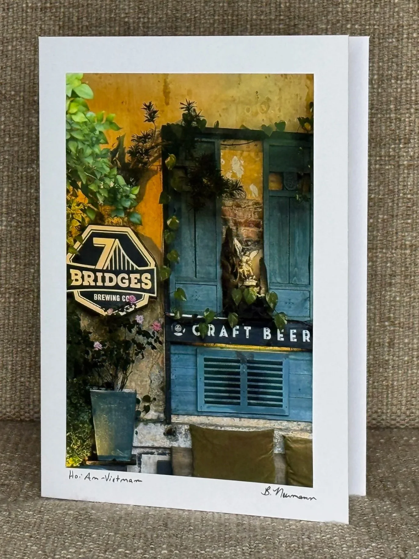Photo of a printed image showing a rustic blue wooden door with greenery in a pot nearby, a sign reading "Bridges Brewing Co," and the words "Craft Beer" on a sign, with the handwritten note "Hoi An, Vietnam" at the bottom.