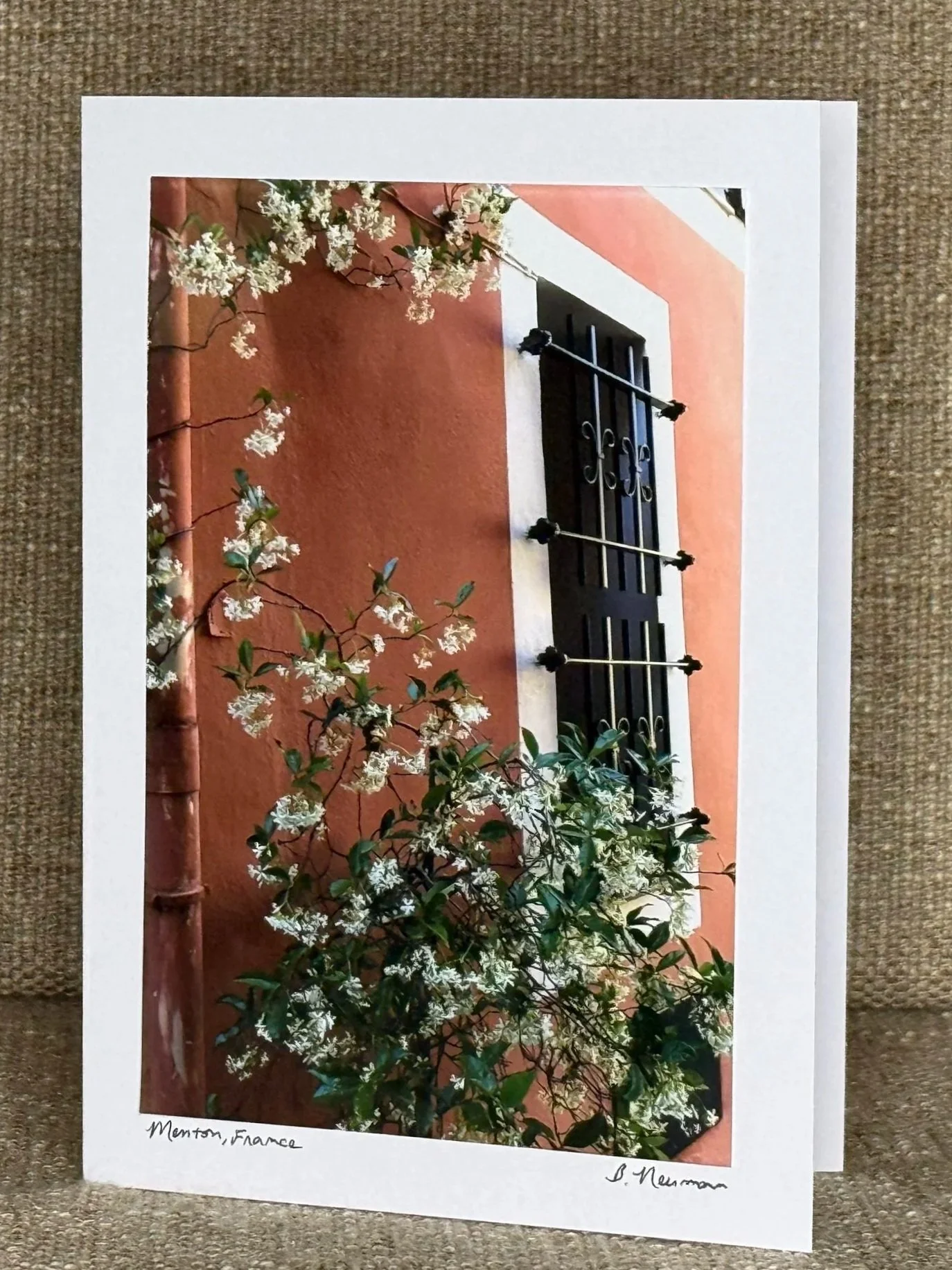 A photo of a corner with light pink walls, a black window with decorative bars, and flowering green bushes in the foreground. The photo is printed on photo paper with handwritten text at the bottom left reading 'Monson, France' and at the bottom righ