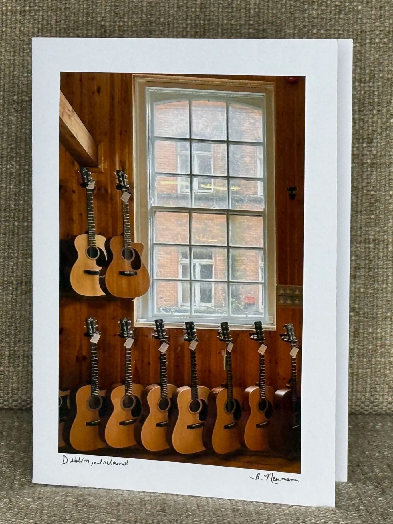 Photograph of a display of multiple guitars on a wooden wall, with a large window showing outside buildings. Handwritten notes at the bottom read 'Dublin, Ireland' and a signature.