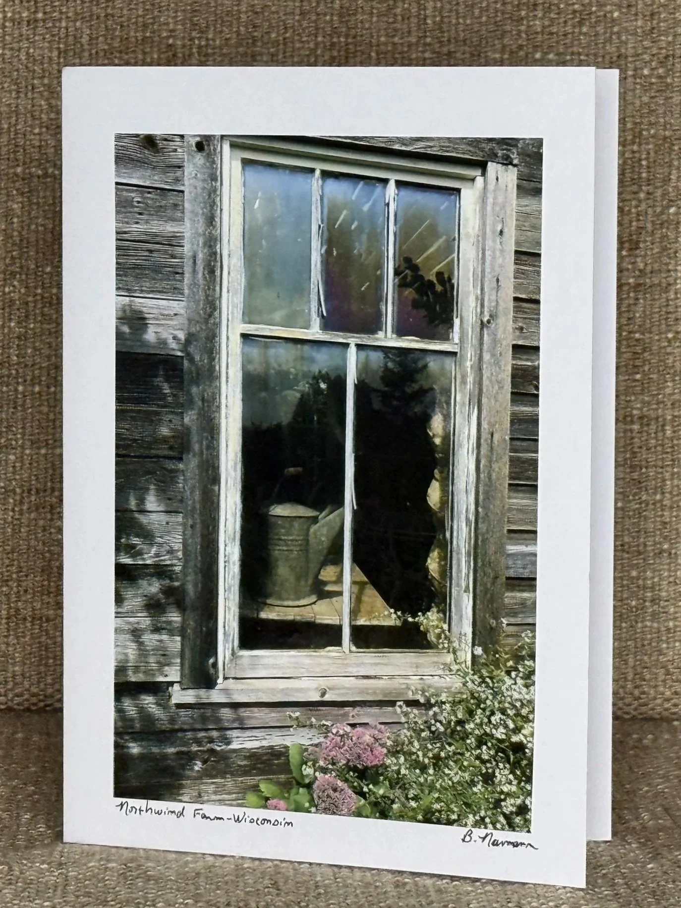 Photograph of a rustic weathered wooden window with four panes, showing a reflection of a person and trees outside, positioned on a wooden building with outdoor plants at the bottom. Handwritten text at the bottom reads 'Northwind Farm-Wisconsin' and