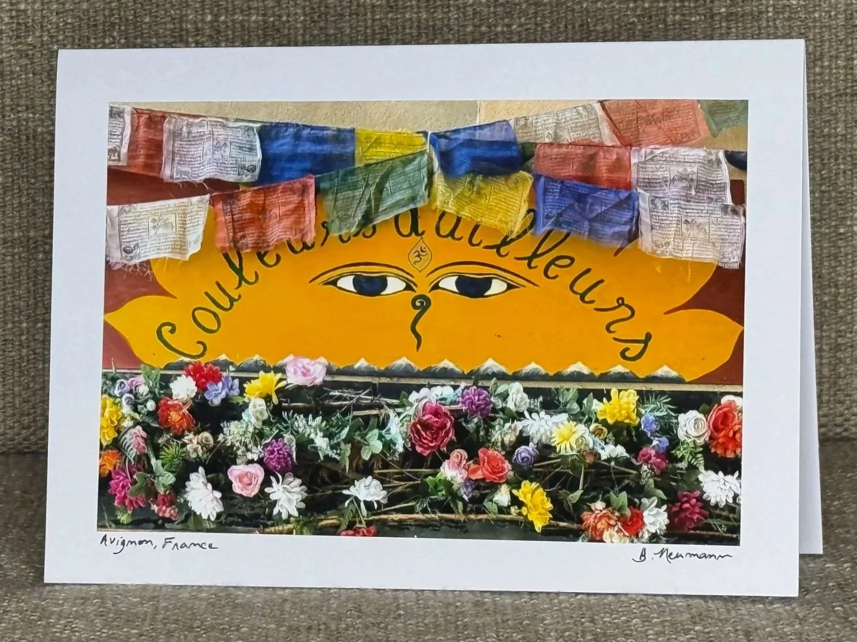 Colorful artwork featuring an eye with a tear drop, surrounded by yellow and orange leaves and a border of various flowers; includes flags and a scarf at the top, with a white border containing handwritten notes at the bottom.