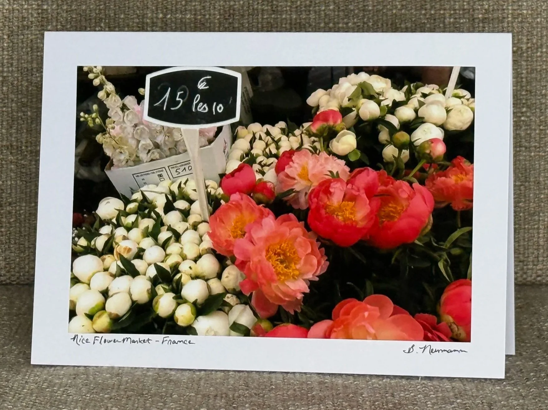 Photo of assorted flowers at a market, priced at 15 Euros for 10, including white and pink blossoms. The photo is framed by a white border with handwritten notes at the bottom indicating it was taken at Nice Flower Market in France, signed by B. Neumann.