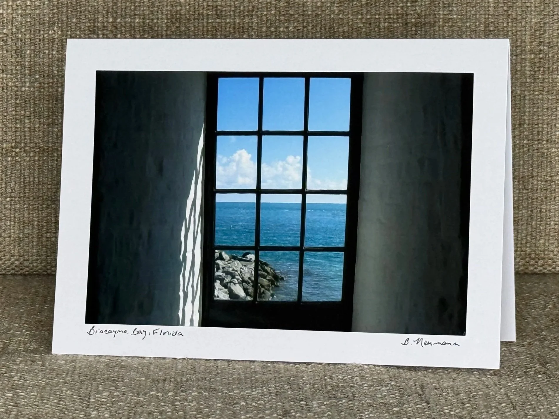 Photograph of a window view of the ocean with rocky shoreline, blue sky, and clouds, framed in a dark structure, with a handwritten note at the bottom reading "Biocayne Bay, Florida".