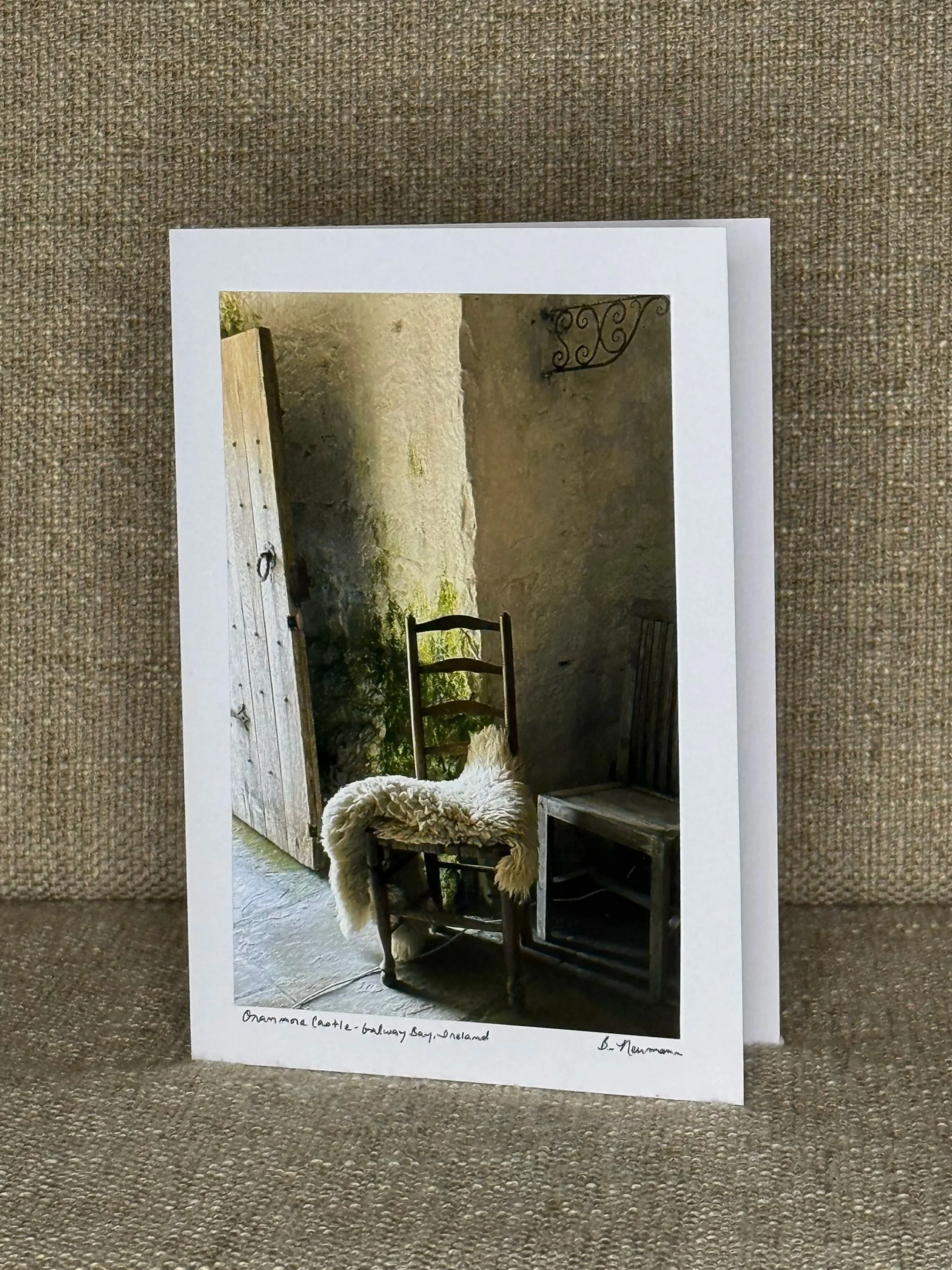 Photo of a printed image on paper showing a rustic scene with a wooden door, an iron wall decoration, and a wooden chair with a sheepskin throw, labeled as 'Onamannan Castle - Ballymena Bay, Ireland'.