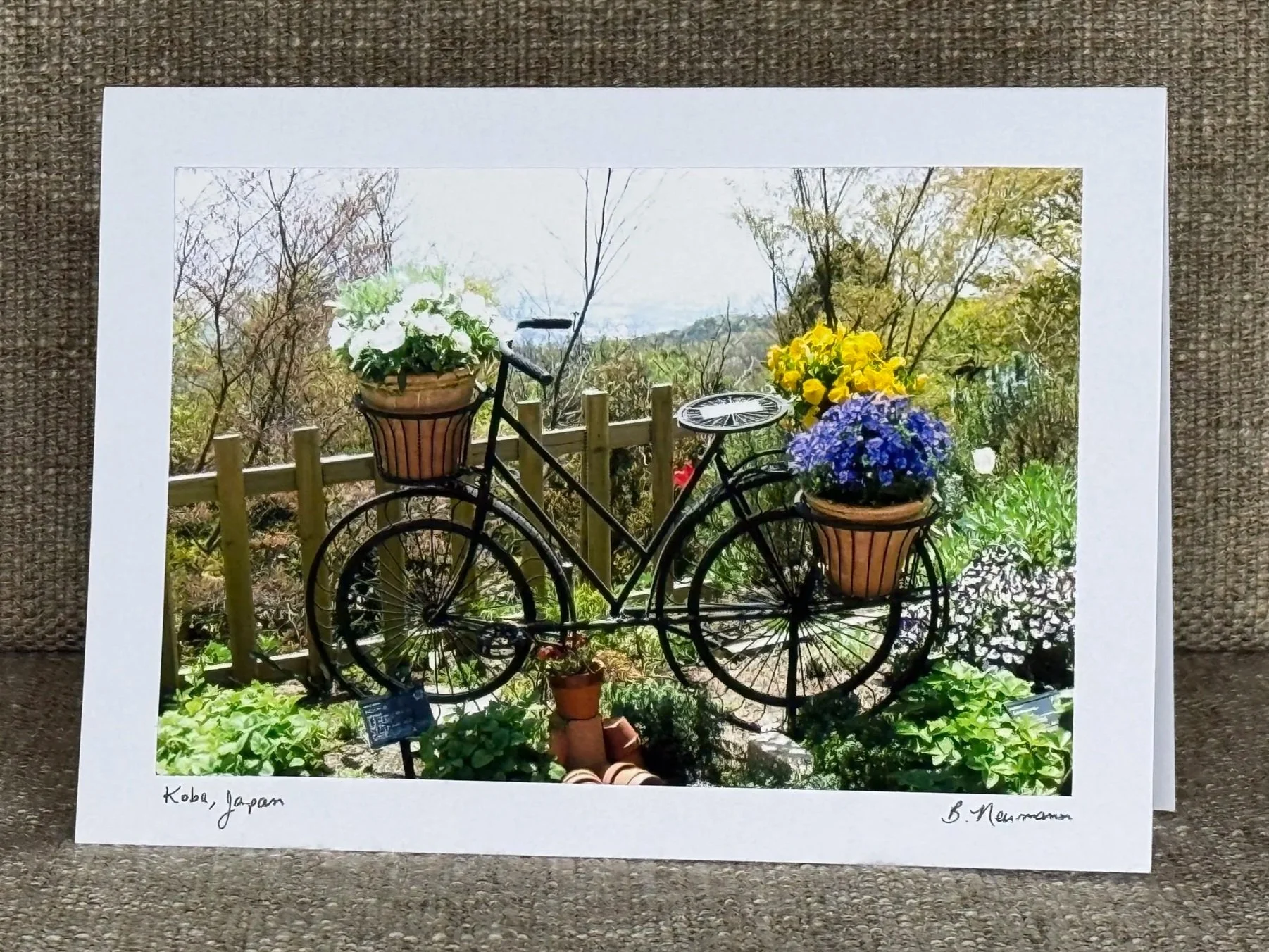 Photograph of a decorative bicycle sculpture with flower pots attached to its basket and seat, located outdoors with trees and a wooden fence in the background, in Kobe, Japan.