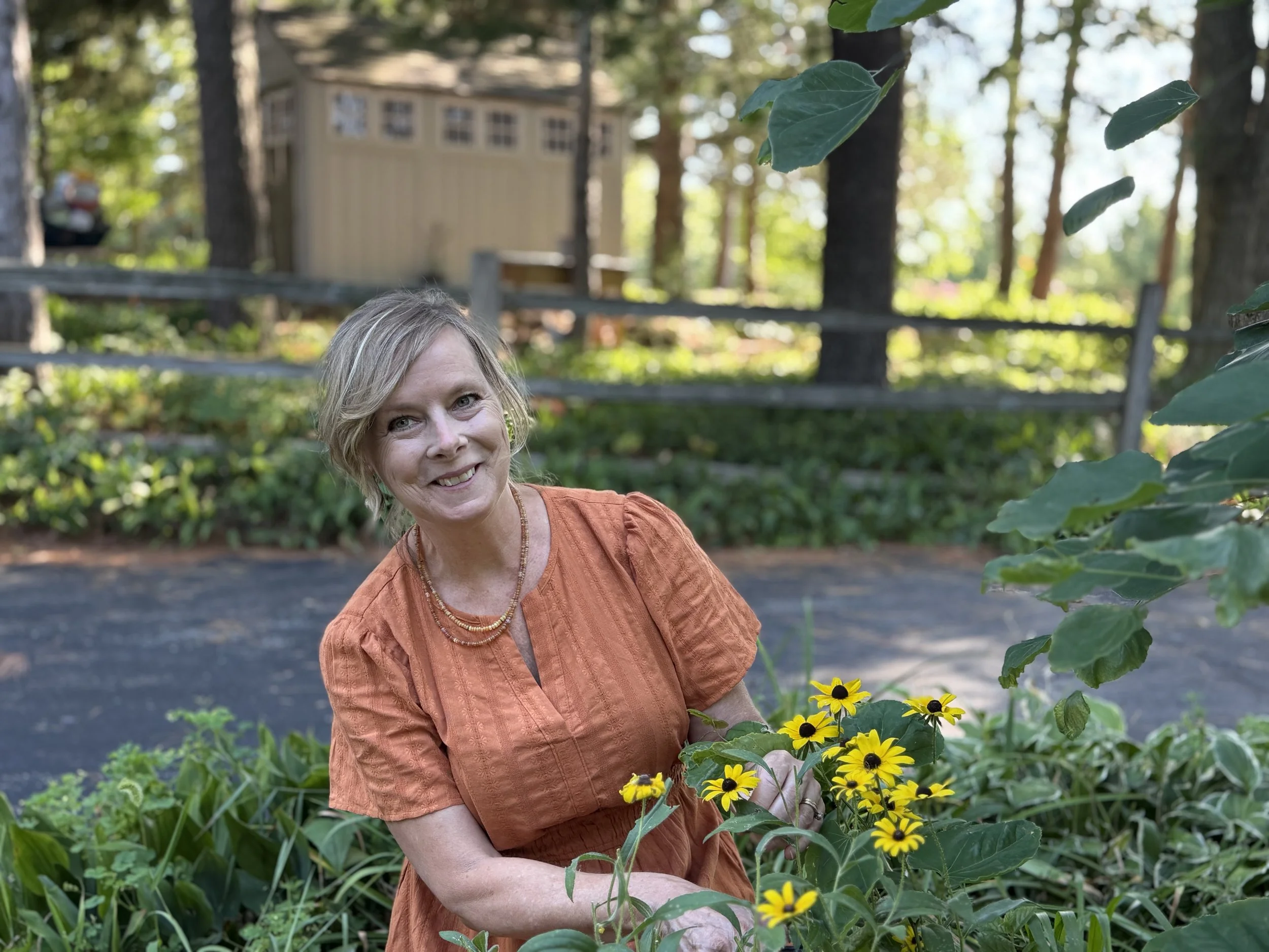 A woman with short blonde hair smiling in a garden, surrounded by yellow flowers and green foliage.