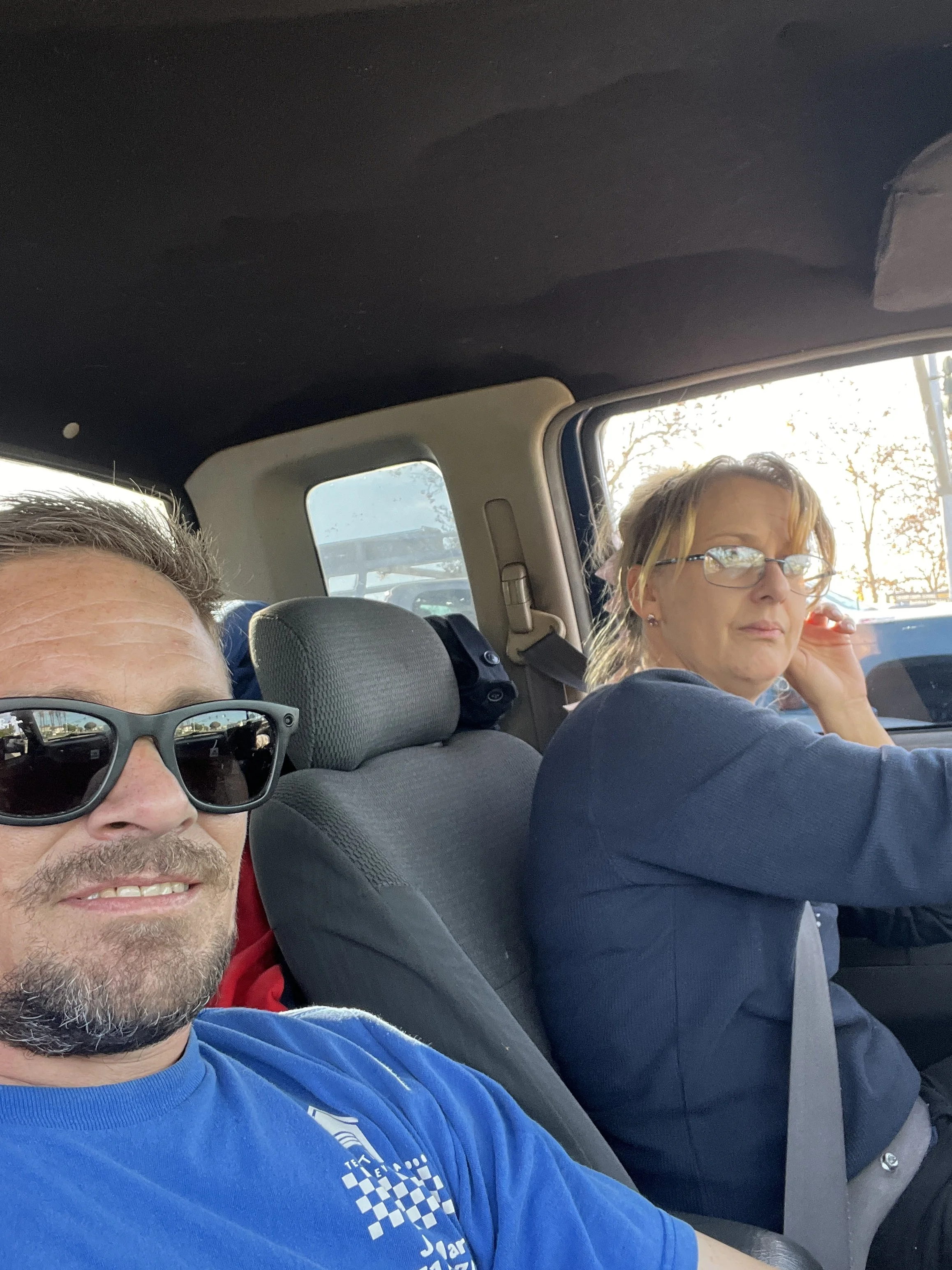 A man with glasses and a beard taking a selfie in a car, seated next to a woman with glasses and blonde hair, talking on a cell phone, with part of the car interior and sunlight visible through the windows.