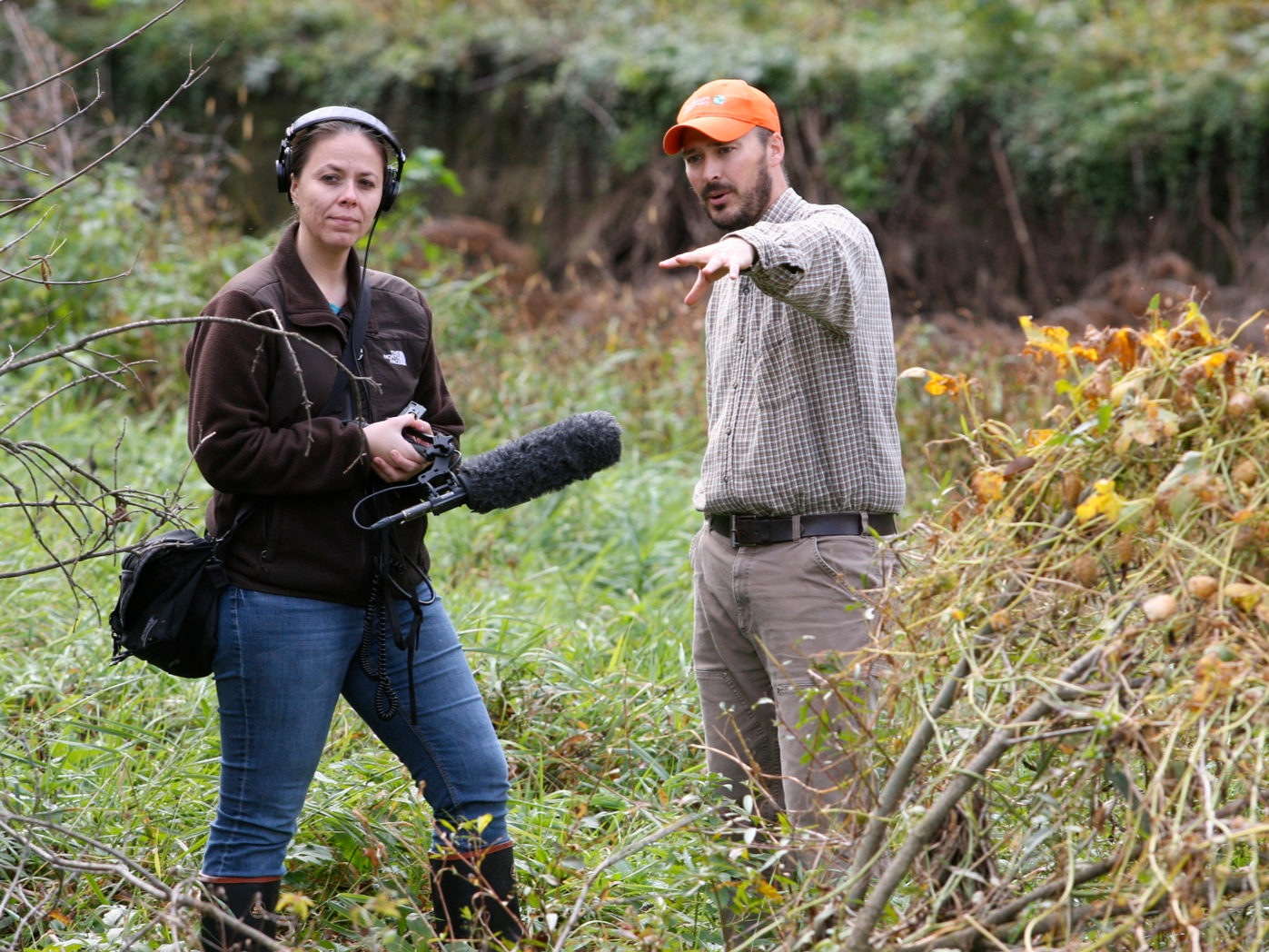 Two people in a forest field, one woman with headphones and a microphone, the man in a checkered shirt and orange hat, gesturing as they talk.