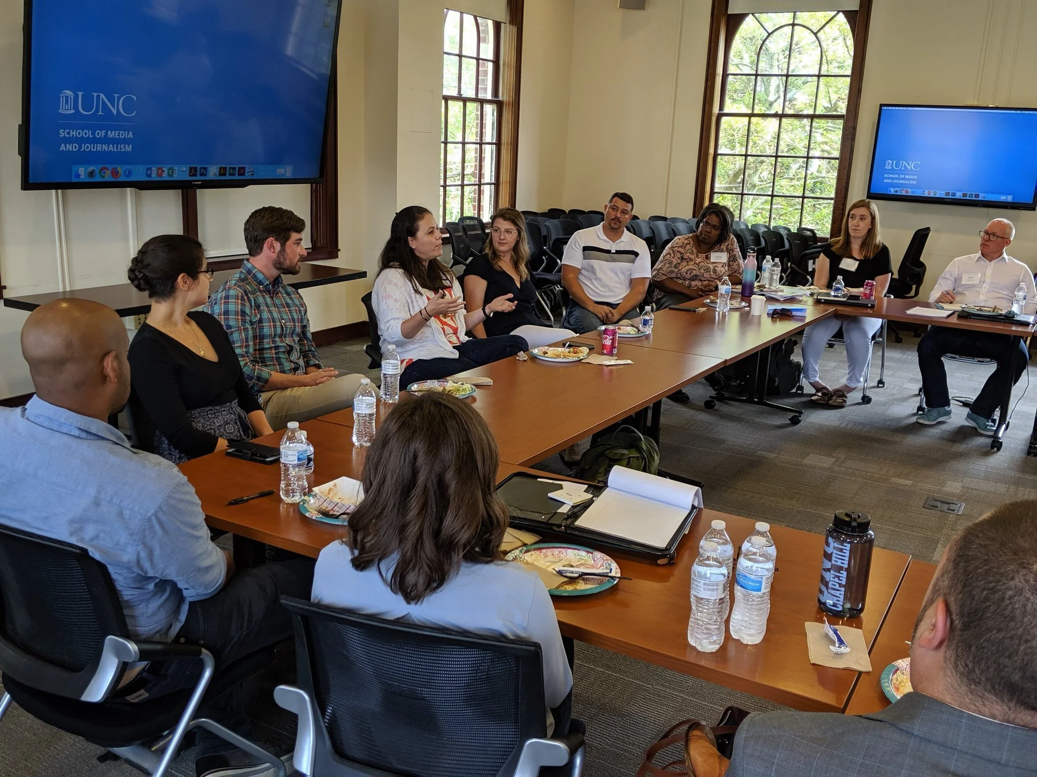 People sitting around a conference table in a room with large windows, having a discussion. Two large screens display the UNC School of Media and Journalism logo.