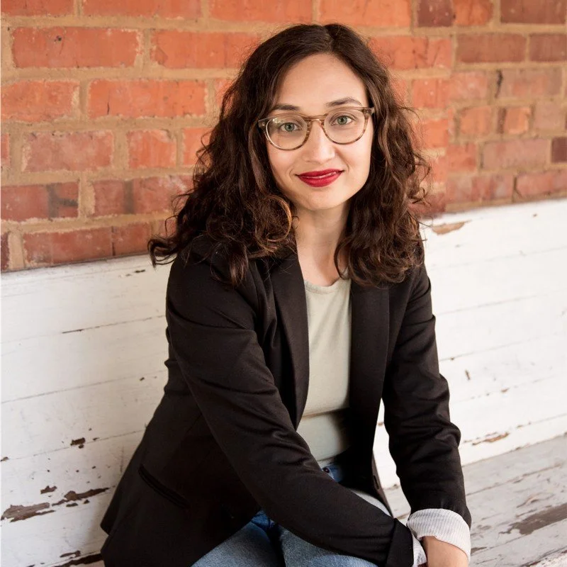 A woman with curly brown hair and glasses, wearing a black blazer and light-colored shirt, sitting on a white wooden bench against a brick wall background.
