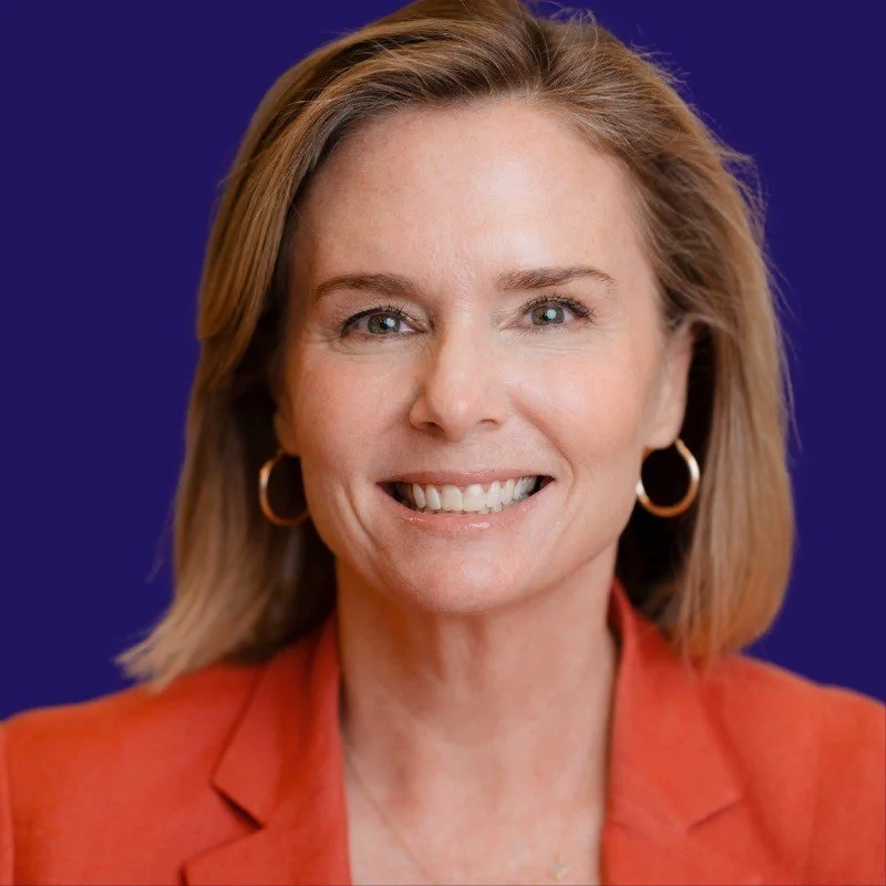 A woman with shoulder-length blonde hair smiling, wearing gold hoop earrings and an orange blazer against a blue background.