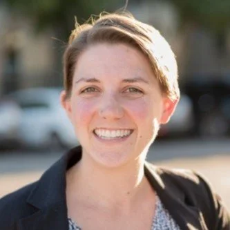 A smiling woman with short hair, wearing a black blazer, outdoors with blurred cars and trees in the background.