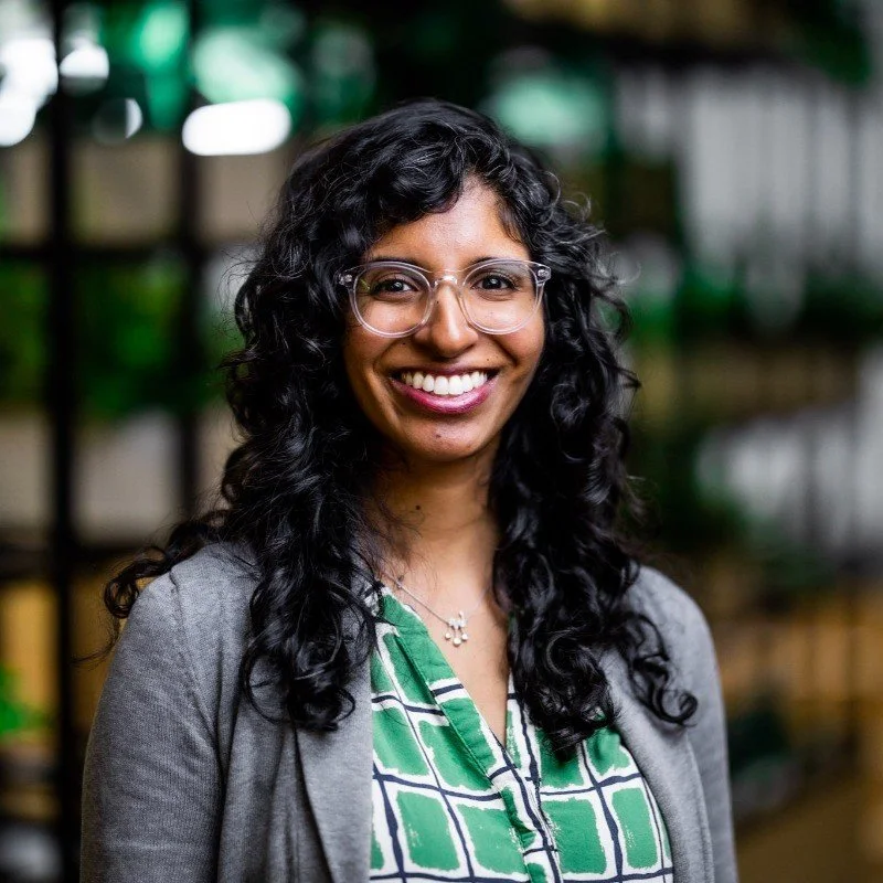 A woman with long, curly black hair, glasses, and a warm smile, standing indoors with a blurred background of green and yellow objects.