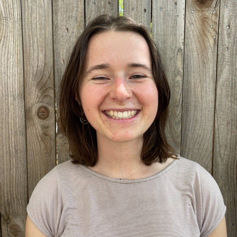 A young woman with short brown hair smiling in front of a wooden fence.