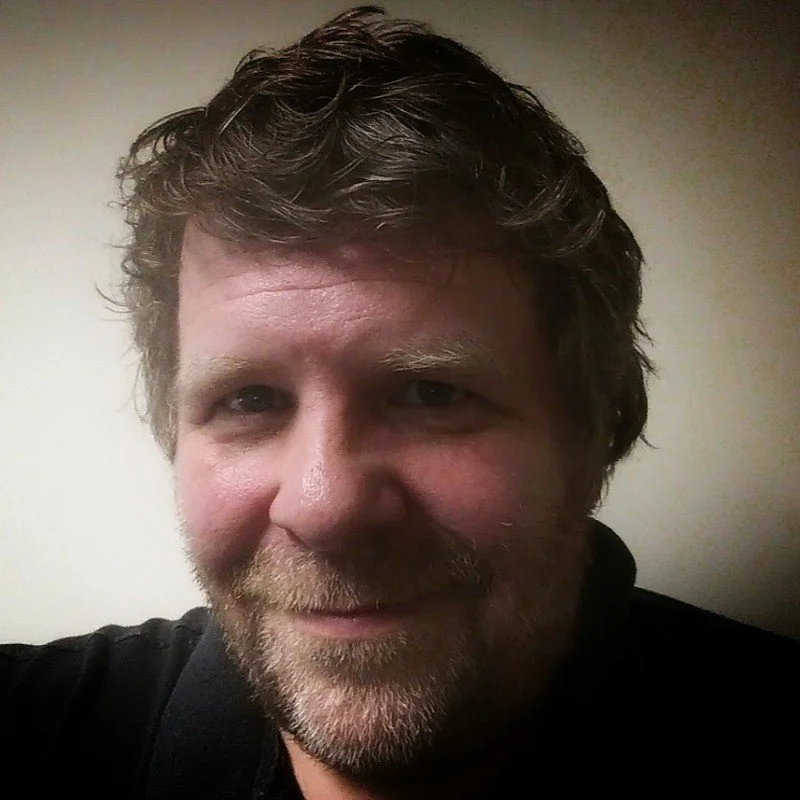 Close-up of a smiling man with curly hair, facial hair, and a black shirt, against a plain background.