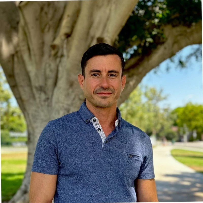 A man with short dark hair and light skin standing outdoors in front of a large tree, wearing a blue collared shirt, with a walkway and greenery in the background.