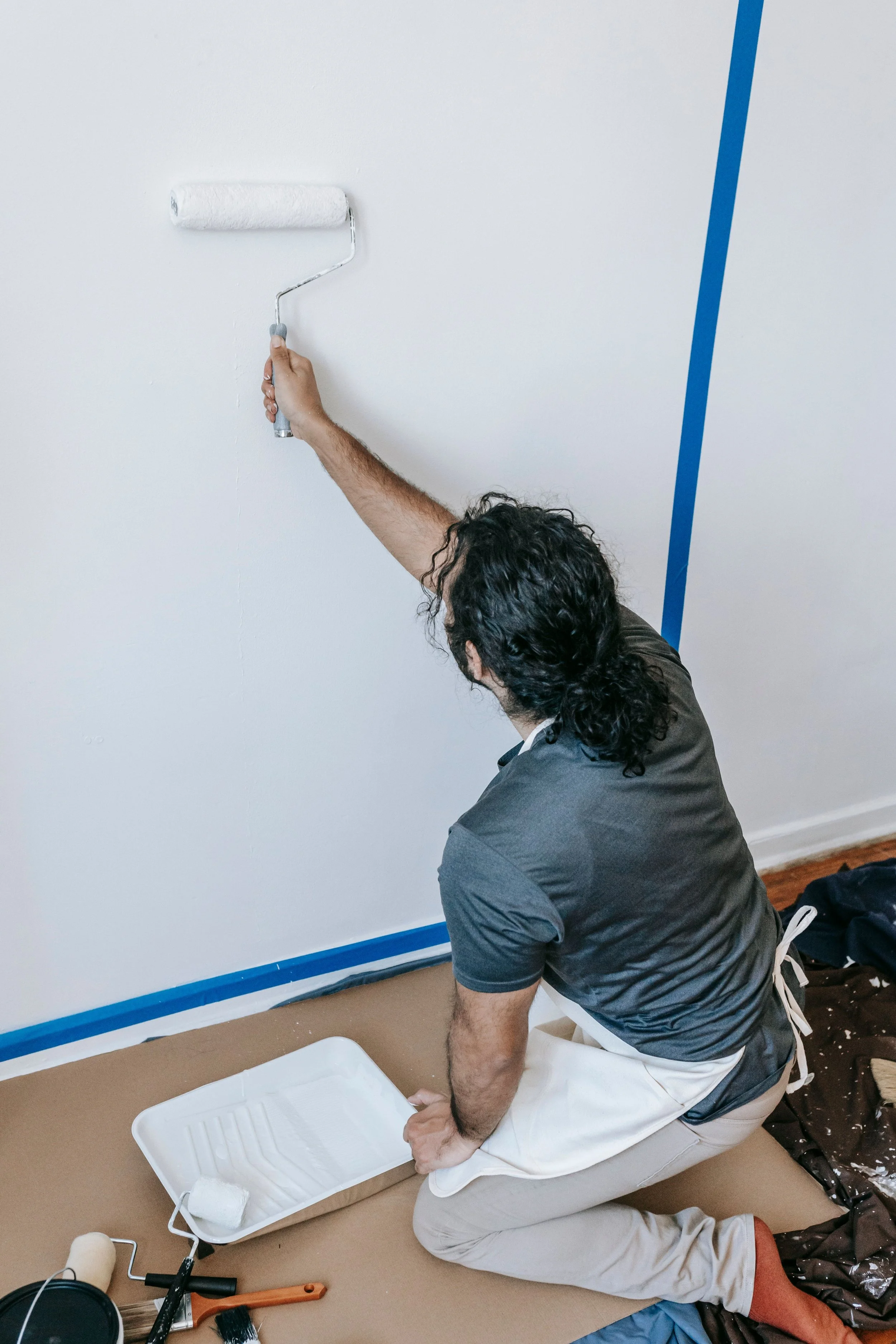 A person crouched down on the floor using a paint roller on a wall. They are wearing a gray shirt, white apron, and beige pants. Painting supplies, including a paint tray with white paint, are nearby on the floor. Blue painter's tape lines the bottom of the wall.