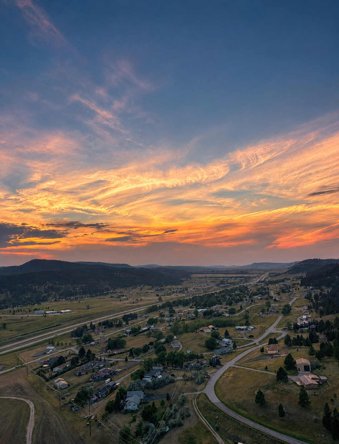 Aerial view of a rural neighborhood at sunset, with winding roads, houses, trees, and distant hills under a colorful sky.