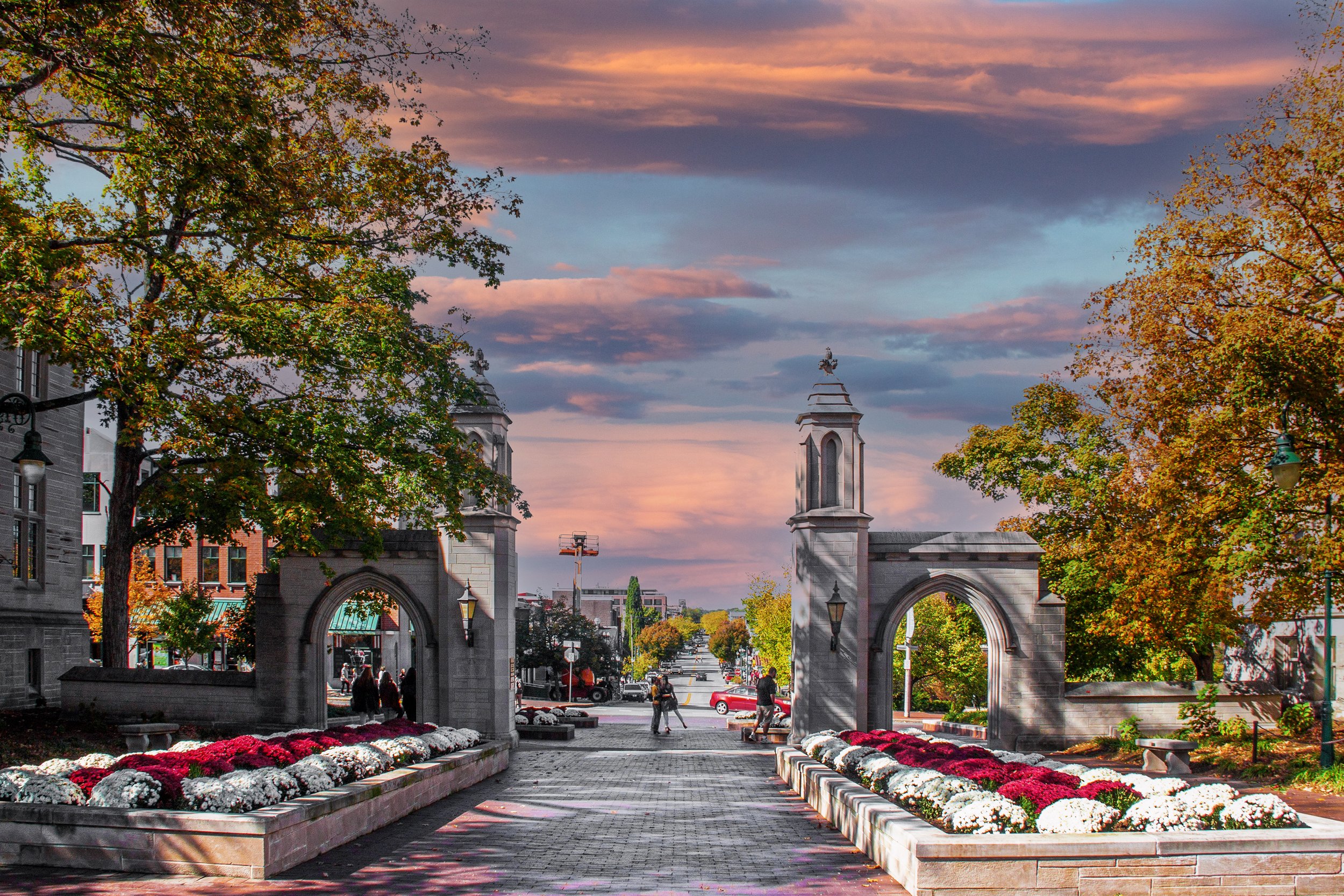 A city scene during sunset with a decorative archway and a floral bed in the foreground, trees with autumn foliage, and buildings in the background.