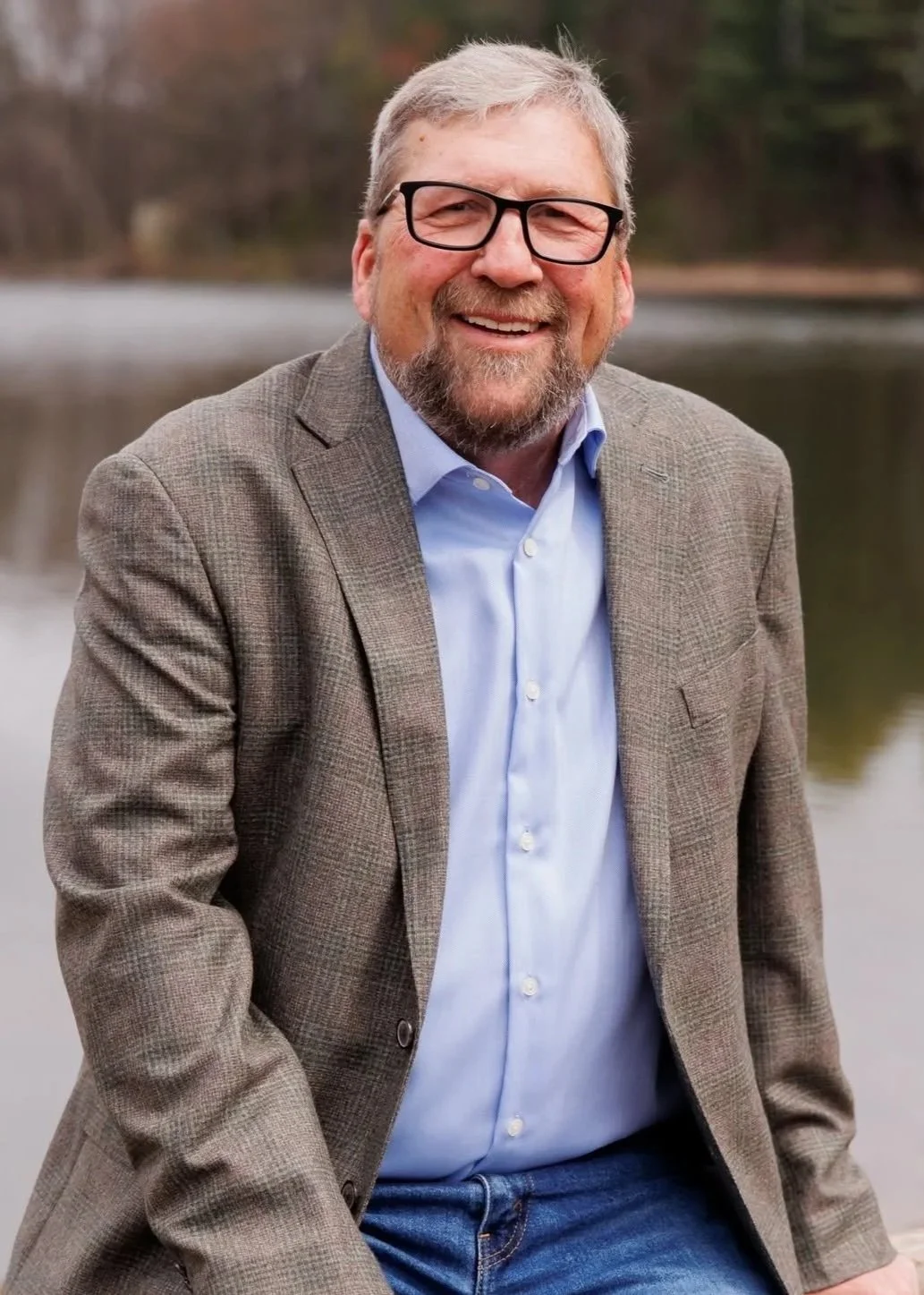 A smiling older man with glasses and a beard, wearing a light blue shirt and a brown blazer, outdoors near a body of water with trees in the background.