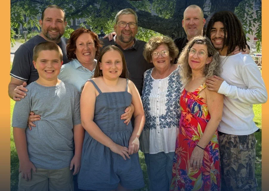 A group photo of nine people, including children and adults, smiling outdoors in a park, with trees and grass in the background