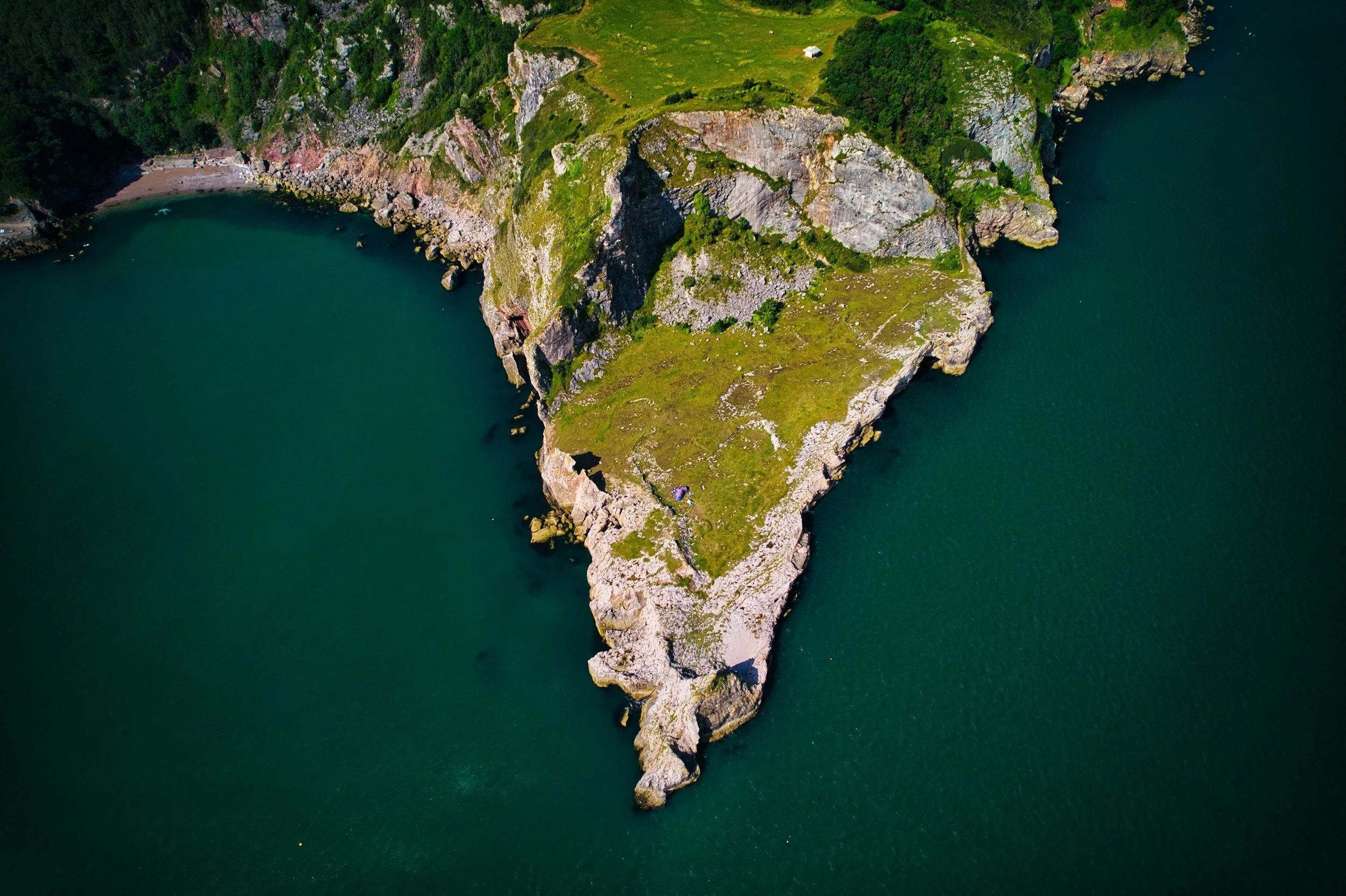 An aerial view of a small, tree-covered island with steep rocky cliffs, surrounded by water. The island features grassy areas, trees, and a small structure near the top.