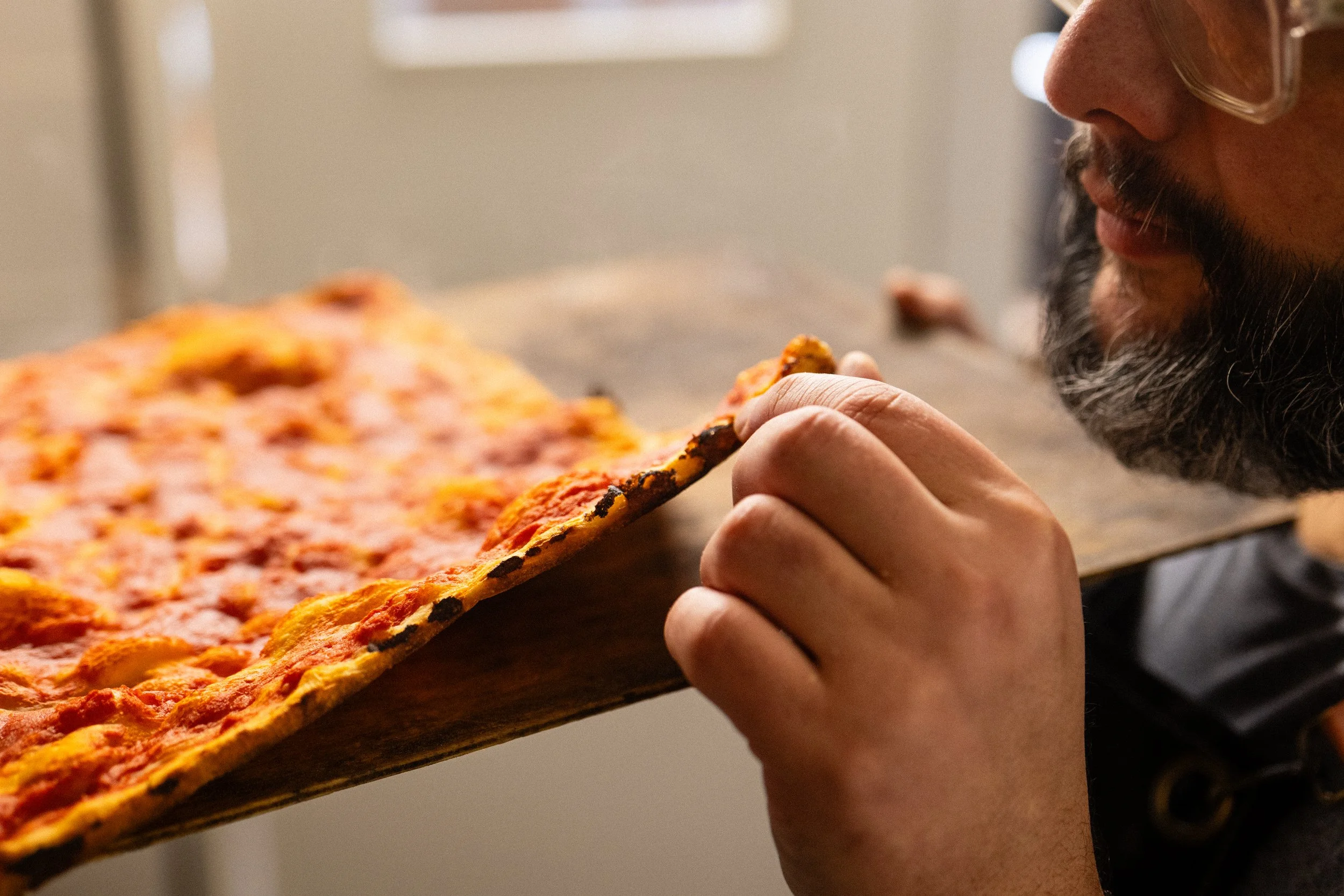 Uomo con barba e occhiali trasparenti che mangia una fetta di pizza con superficie croccante e formaggio fuso.