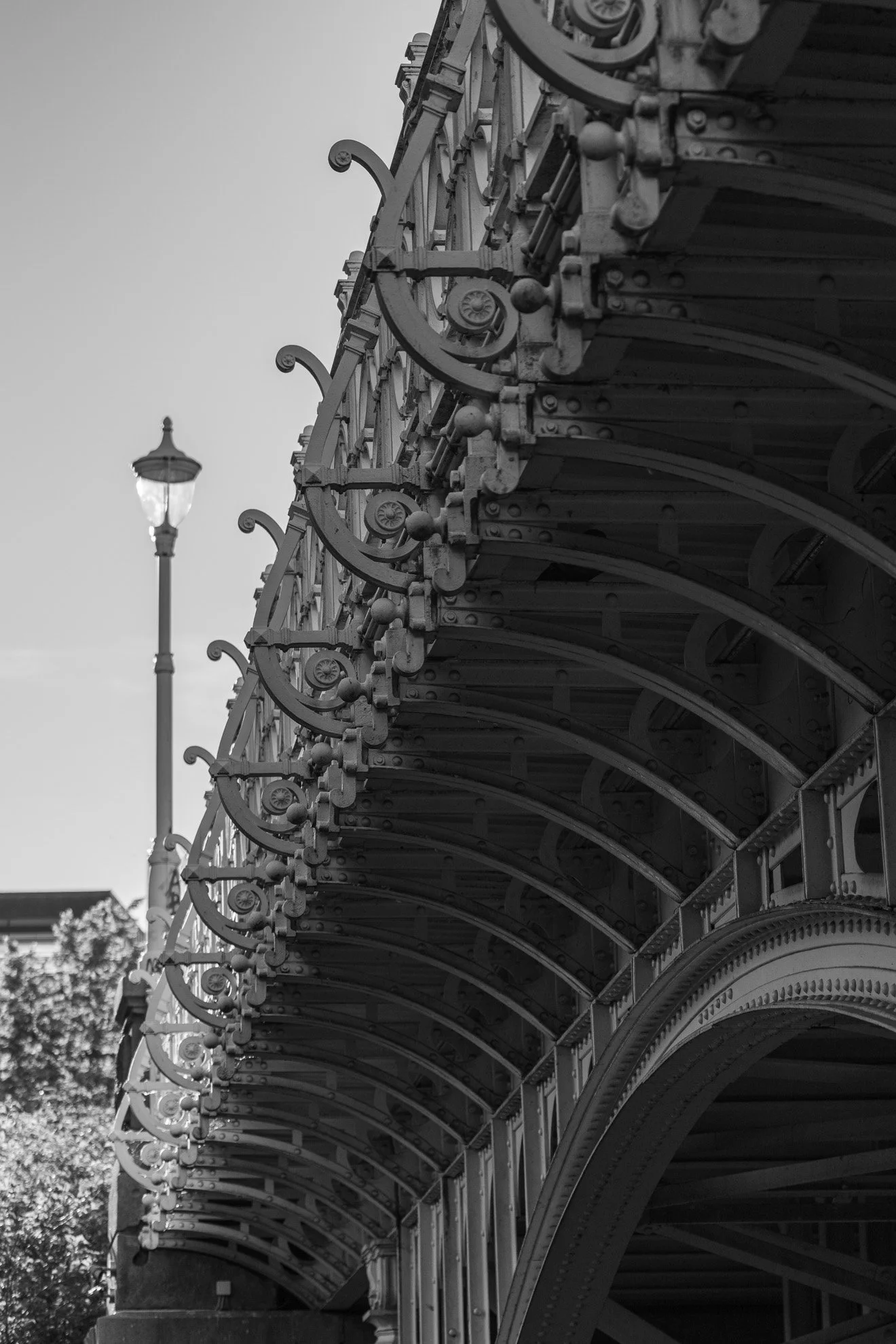 Black and white photo of an ornate iron architecture structure, with a street lamp and trees in the background.
