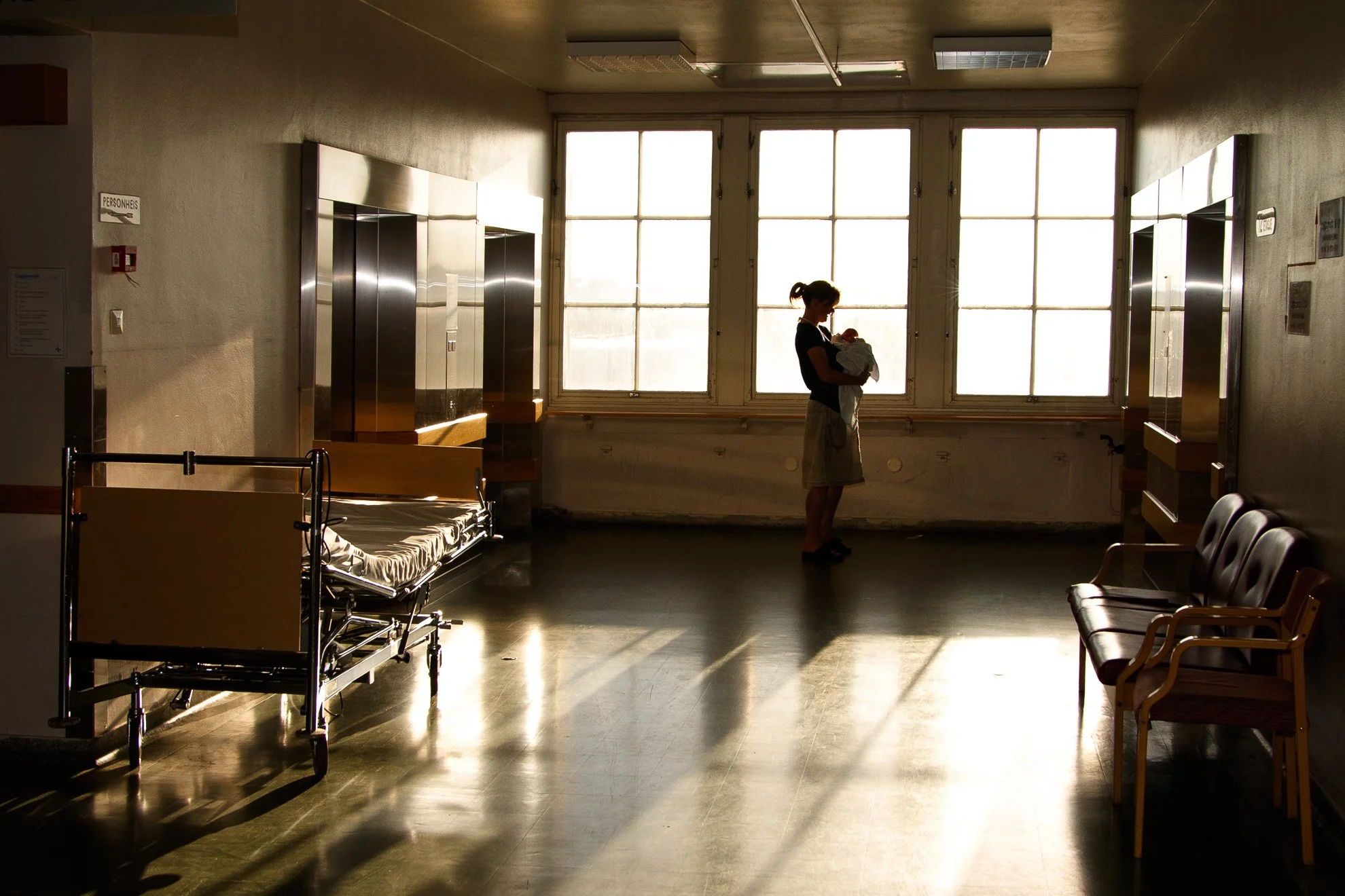 A hospital waiting area with empty chairs and an empty hospital bed, a woman holding a baby near large windows, sunlight illuminating the room.