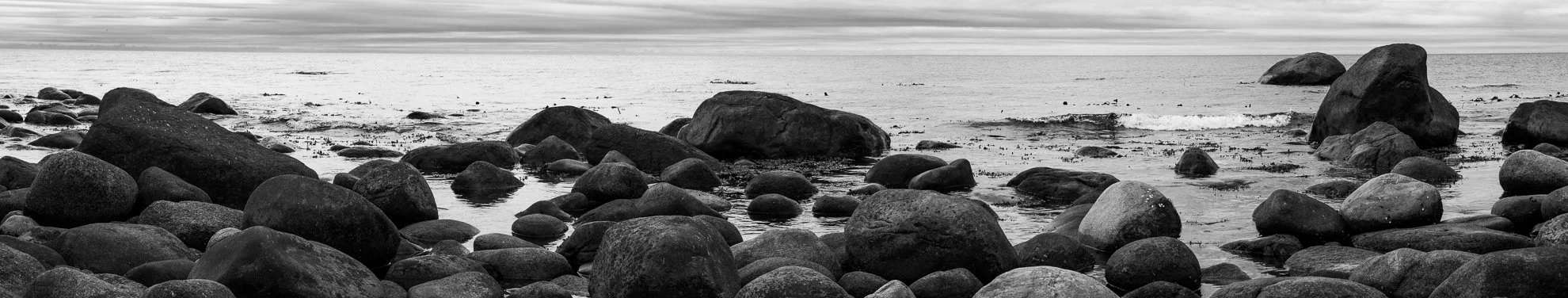 Black and white photo of a rocky shoreline with large smooth boulders scattered along the beach, calm water, and a cloudy sky.