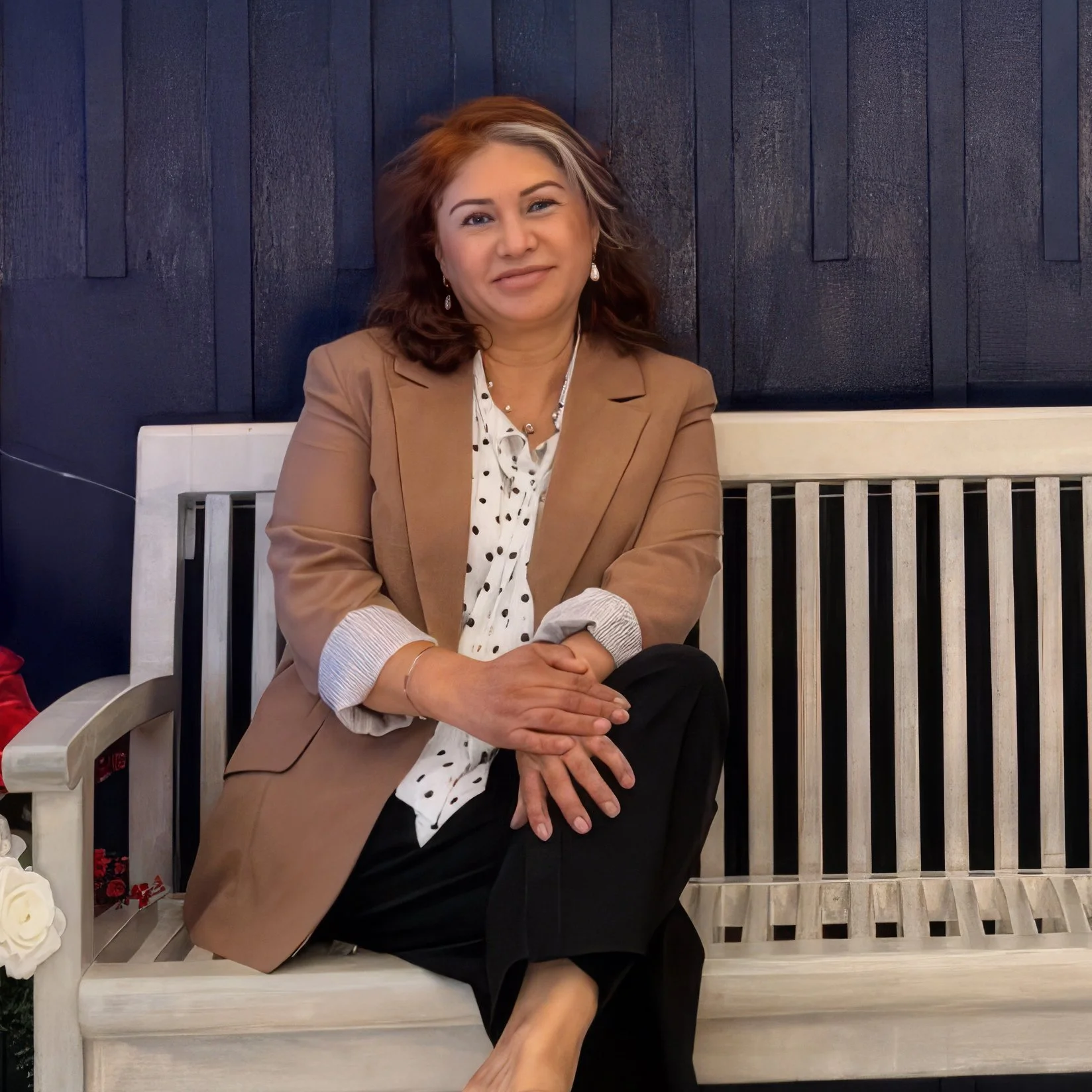 Ana Rivera, in front of a dark blue wooden wall, wearing a white blouse with black polka dots and black pants, smiling with one eye winking.